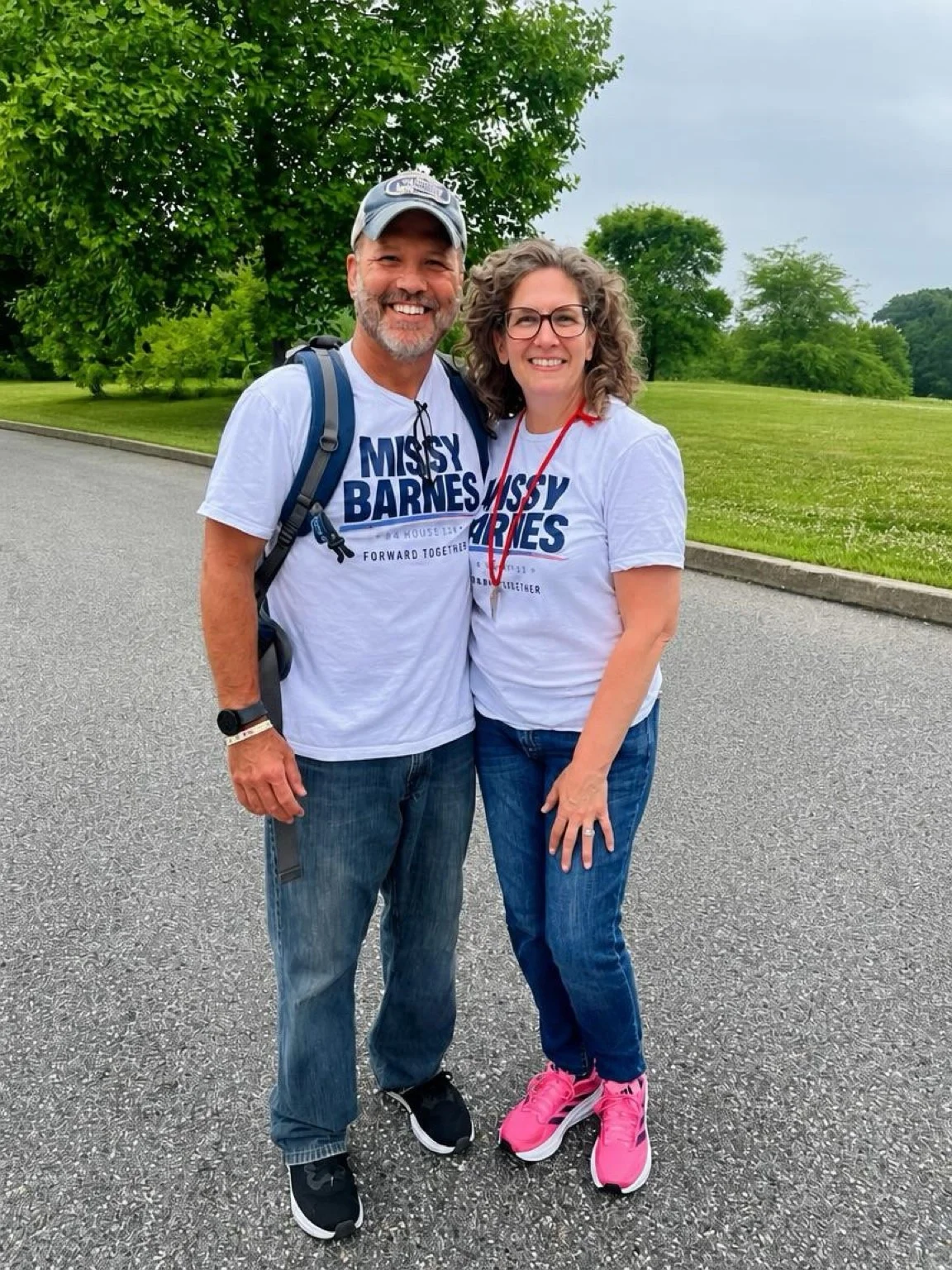 A smiling man and woman standing outside on a paved path, with green trees and grass in the background. They are wearing matching Missy Barnes 4th of July t-shirts. The man has a backpack, a cap, and a smartwatch. The woman has curly hair, glasses, and is wearing pink sneakers.