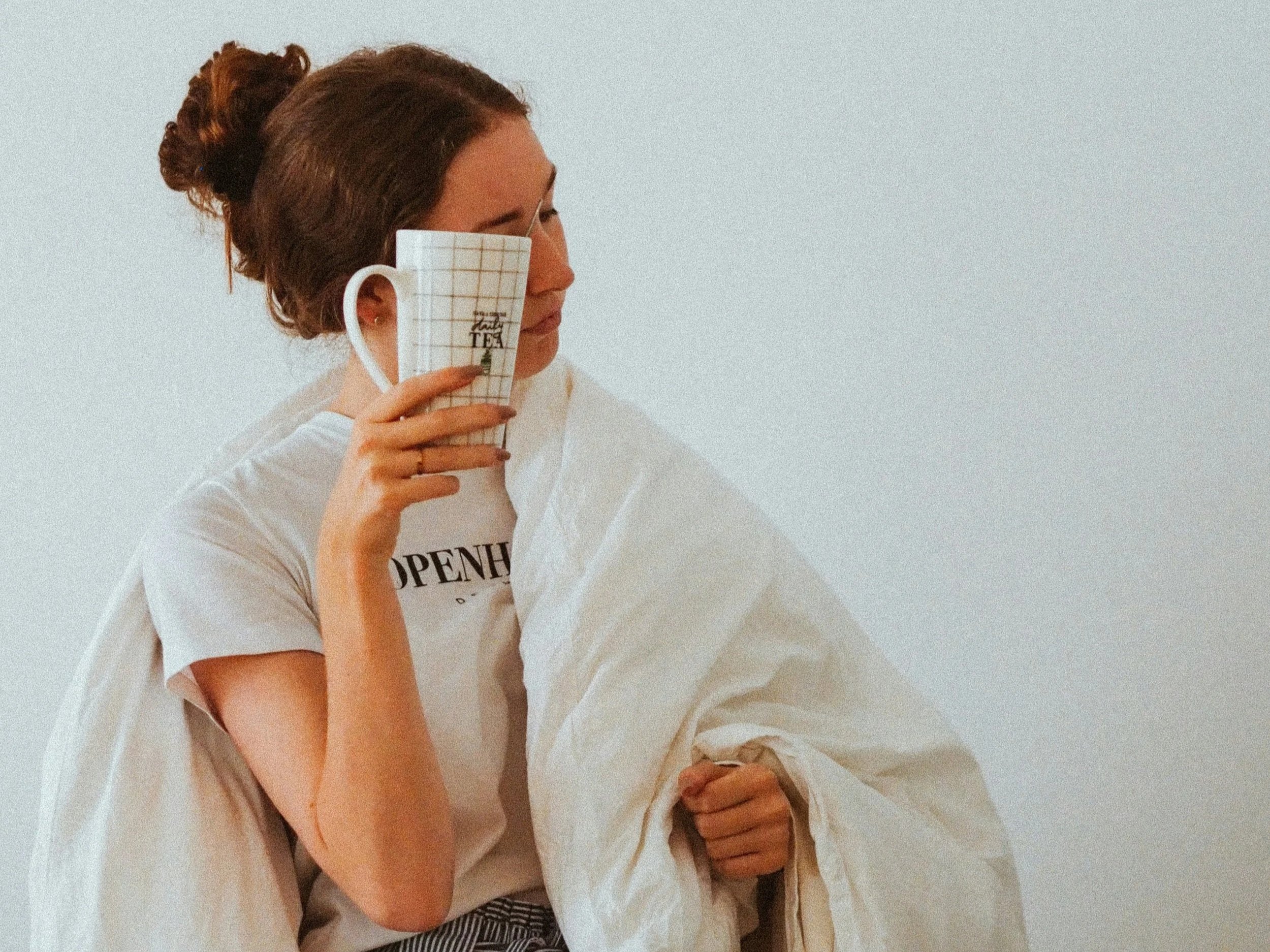 Eine Frau hält sich morgens nach dem Aufwachen eine Teetasse an die Stirn. Das Bild vermittelt Müdigkeit und den Wunsch nach einem ruhigen Start in den Tag.