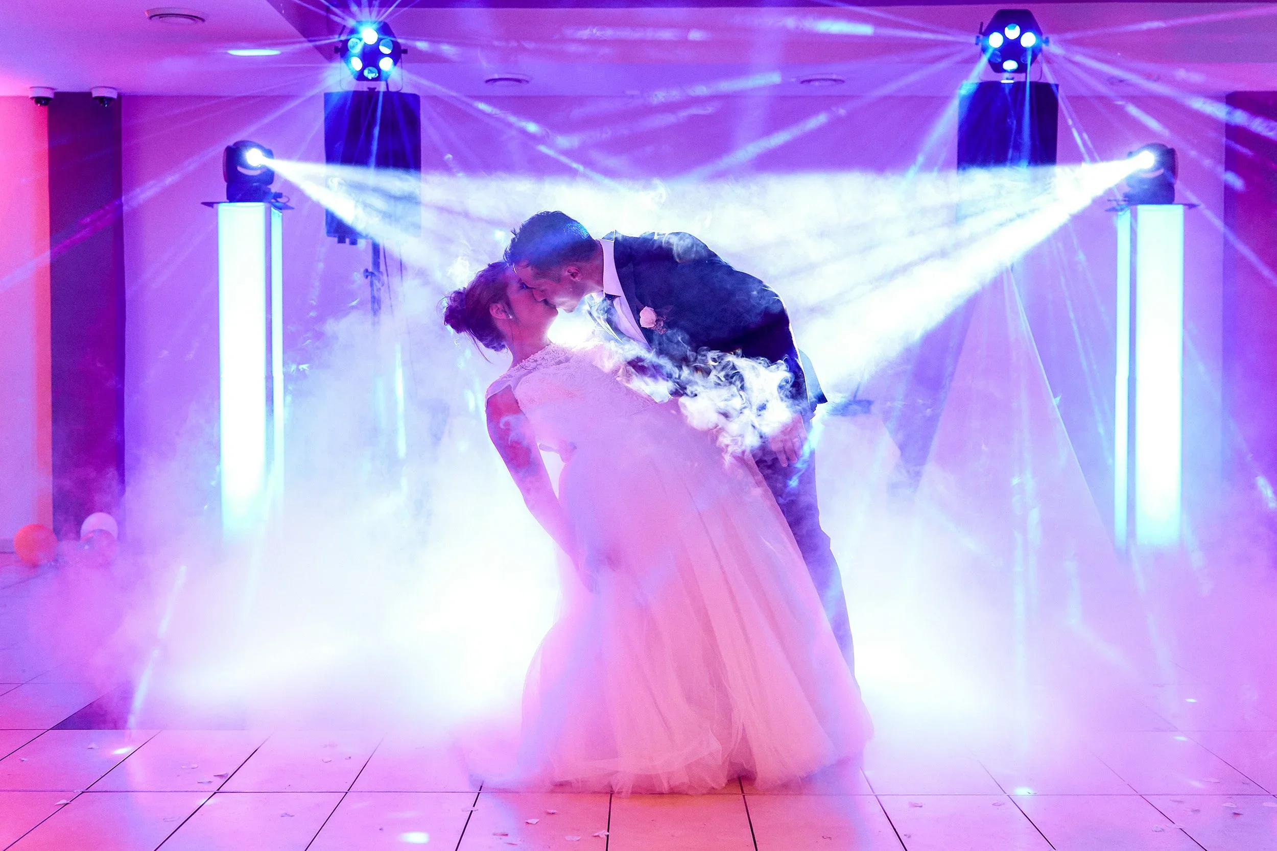 A newlywed couple dancing at their wedding reception. The groom is wearing a dark suit and the bride is in a white wedding gown. Colorful lighting and fog effects create a romantic atmosphere.