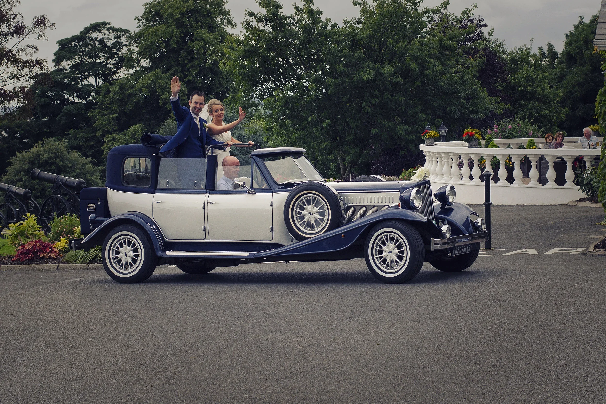 A newlywed couple dressed in wedding attire riding in a vintage convertible car, waving happily. They are driving through a scenic area with lush green trees and a white balcony with colorful flowers in the background. Some guests are seated on the b