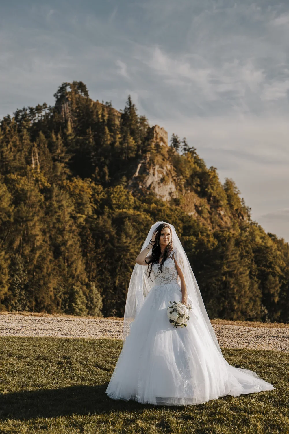 A bride in a white wedding gown holding a bouquet of white flowers stands outdoors on grass with a mountainous, forested backdrop and a partly cloudy sky.