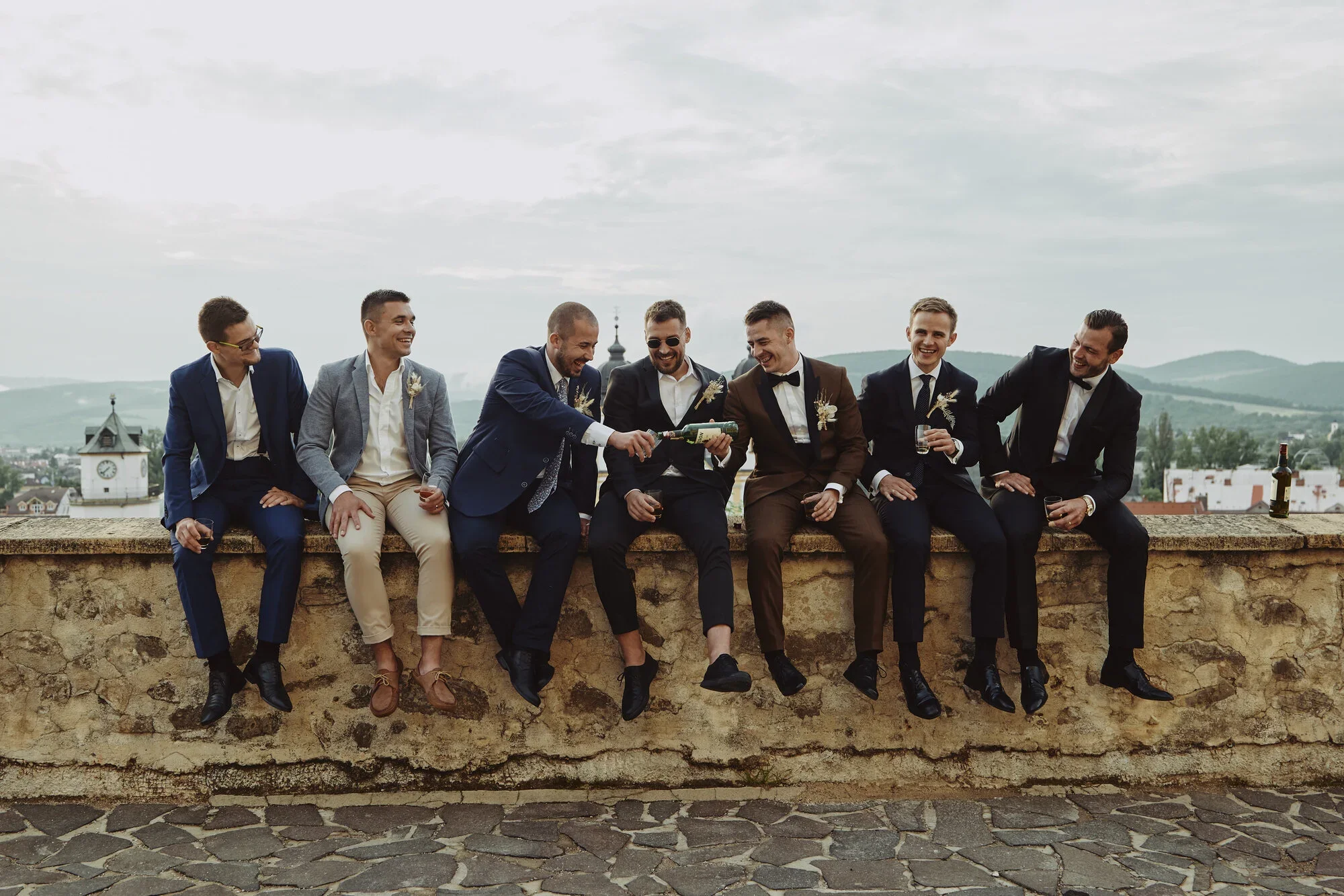 A group of seven men in formal suits sitting on a stone wall outdoors, laughing and socializing, with some holding glasses of drinks and a bottle of champagne, during overcast weather with scenic hills in the background.