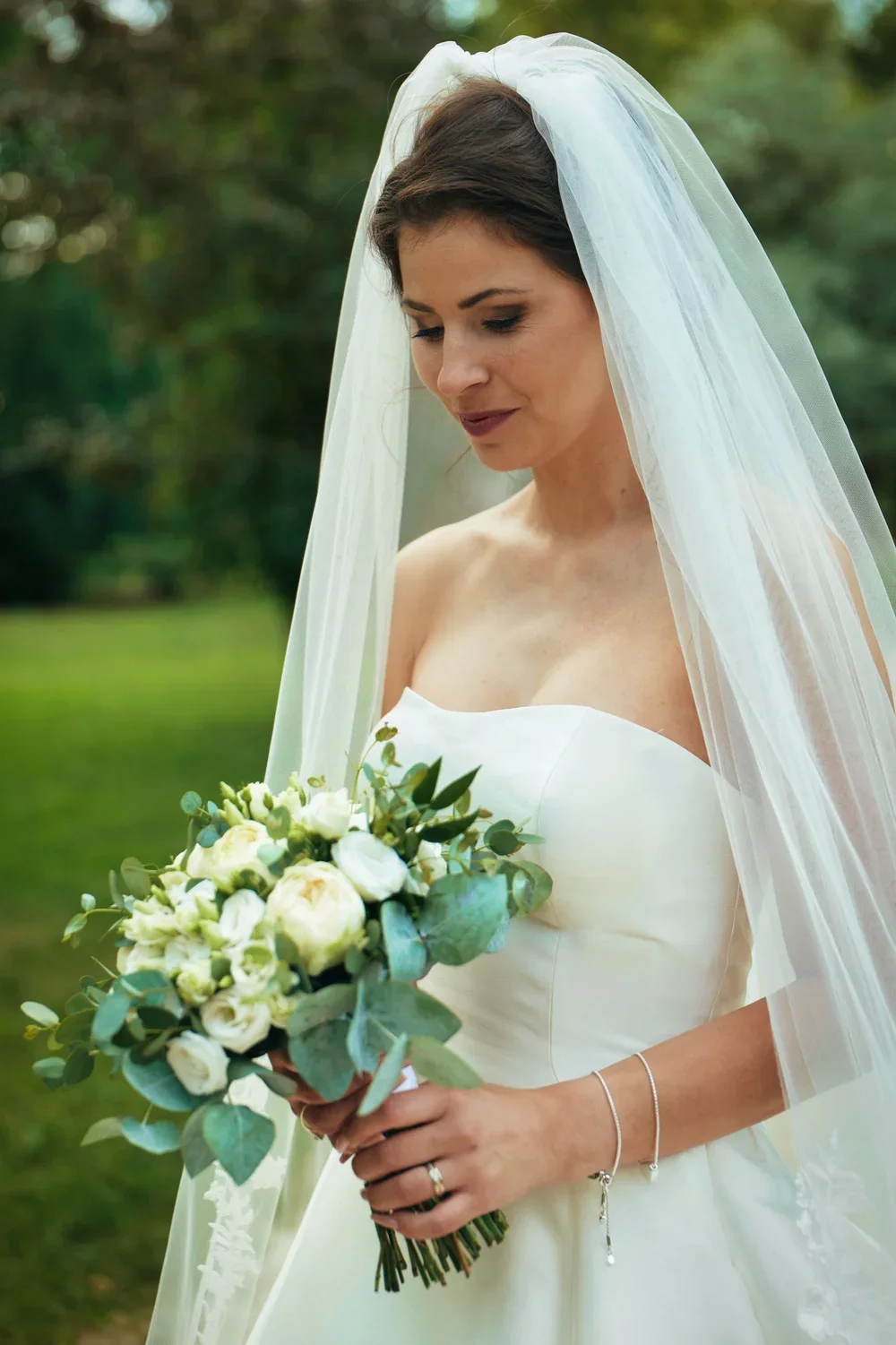 A bride in a strapless wedding dress holding a bouquet of white and green flowers, with a white veil over her dark hair, standing outdoors with green trees in the background.