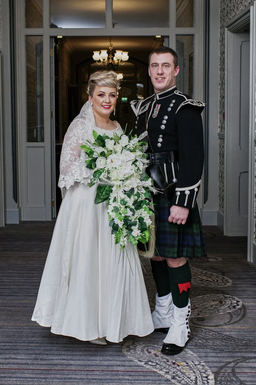 A bride and groom in wedding attire standing inside a decorative hallway. The bride wears a white lace dress and veil, holding a large bouquet of white flowers and greenery. The groom wears a traditional military kilt and jacket, with medals and spat