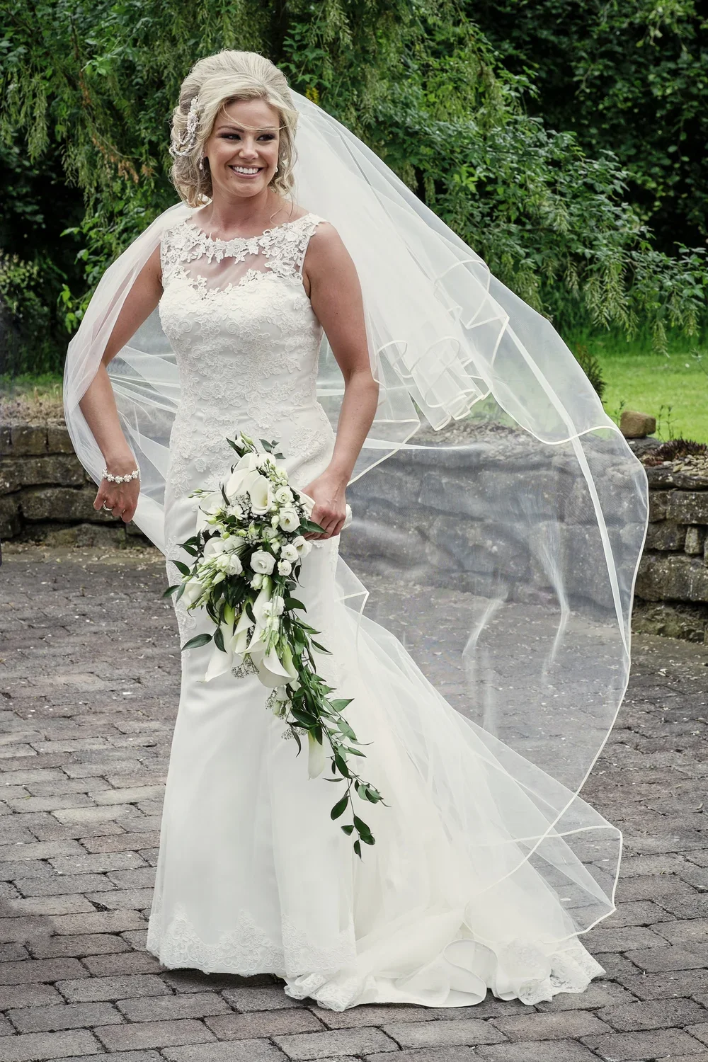 Joyful bride in a white lace wedding gown holding a cascading bouquet of white flowers and greenery, standing outdoors on a cobblestone path with green trees and grass in the background.