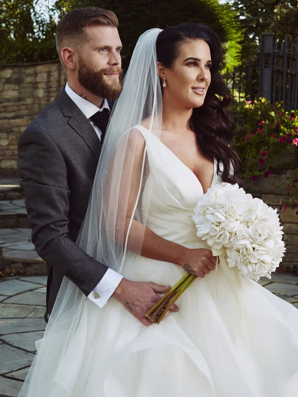 A bride and groom standing outdoors on their wedding day, with the bride holding a bouquet of white flowers and the groom holding her hand, both smiling softly.