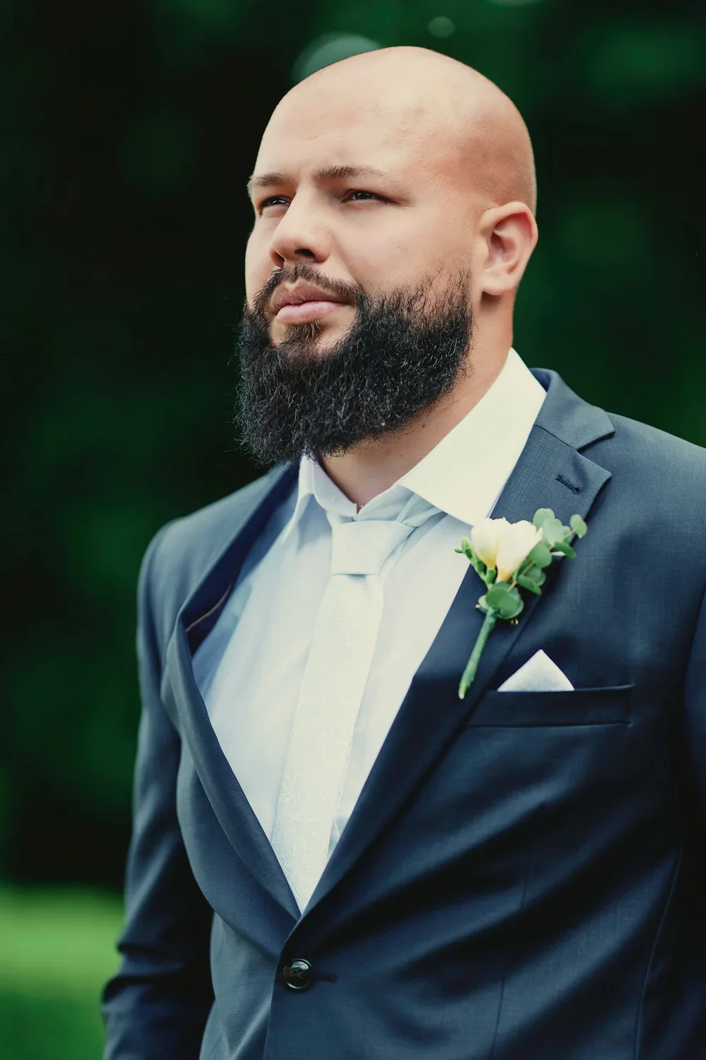 A man dressed in a dark suit, white shirt, and white tie, with a boutonniere on his lapel, standing outdoors with a serious expression.