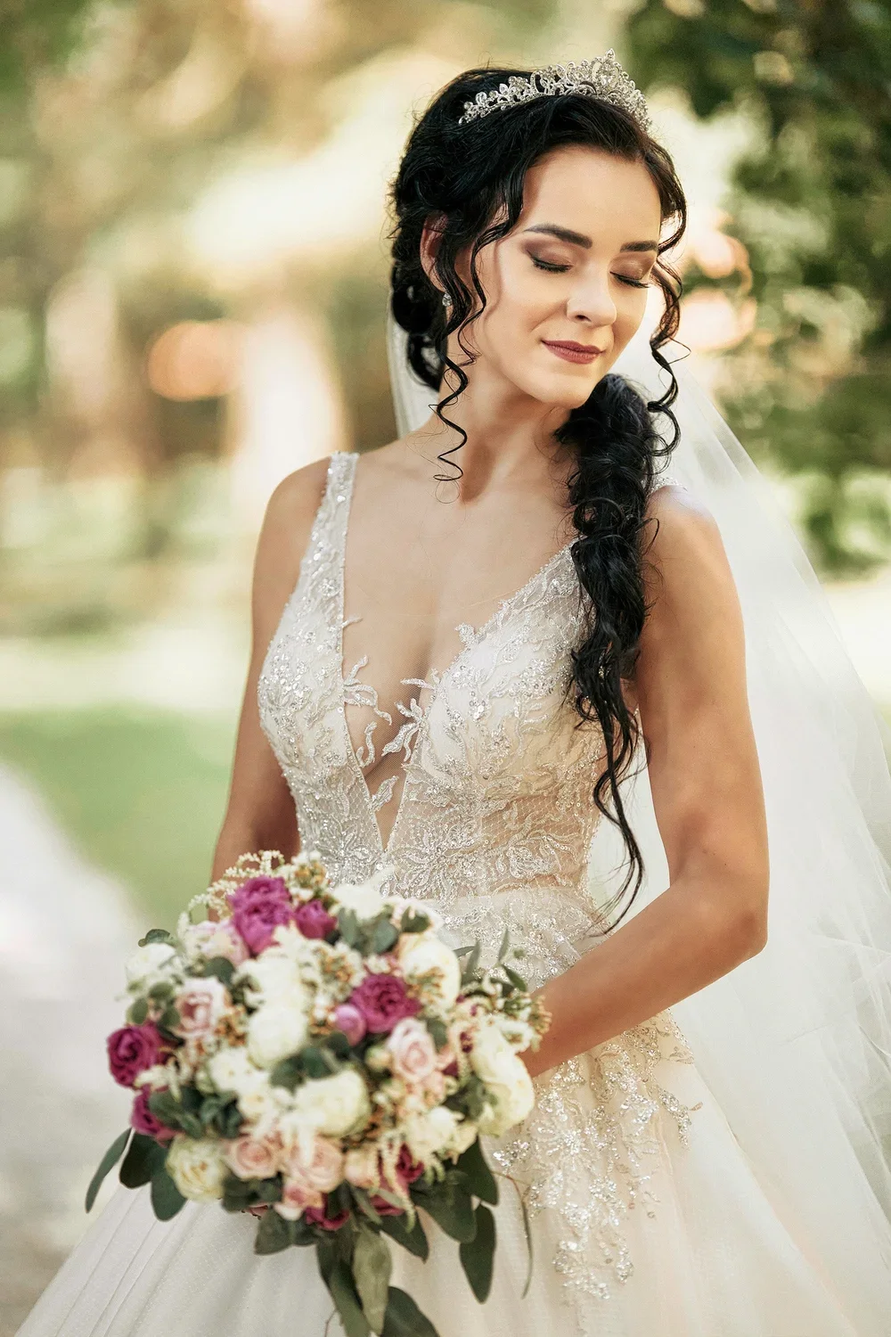 A bride in a wedding gown holding a bouquet of pink and white flowers, with dark hair styled in curls and wearing a tiara and veil.