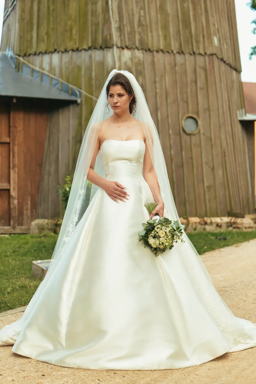 A bride in a white wedding gown holding a bouquet standing outdoors near a wooden building.