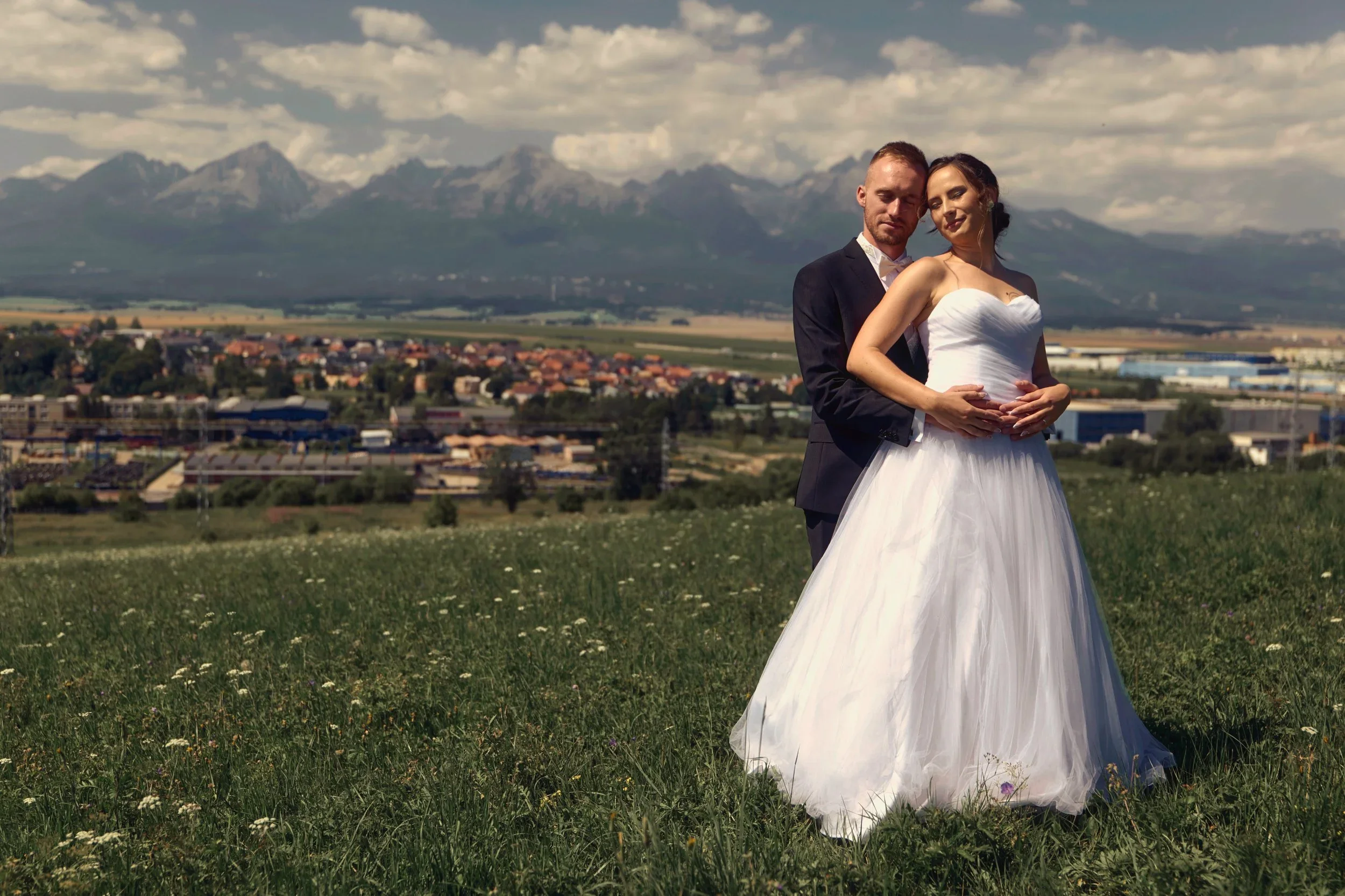 A bride and groom standing in a grassy field with wildflowers, mountains in the background, and a partly cloudy sky.