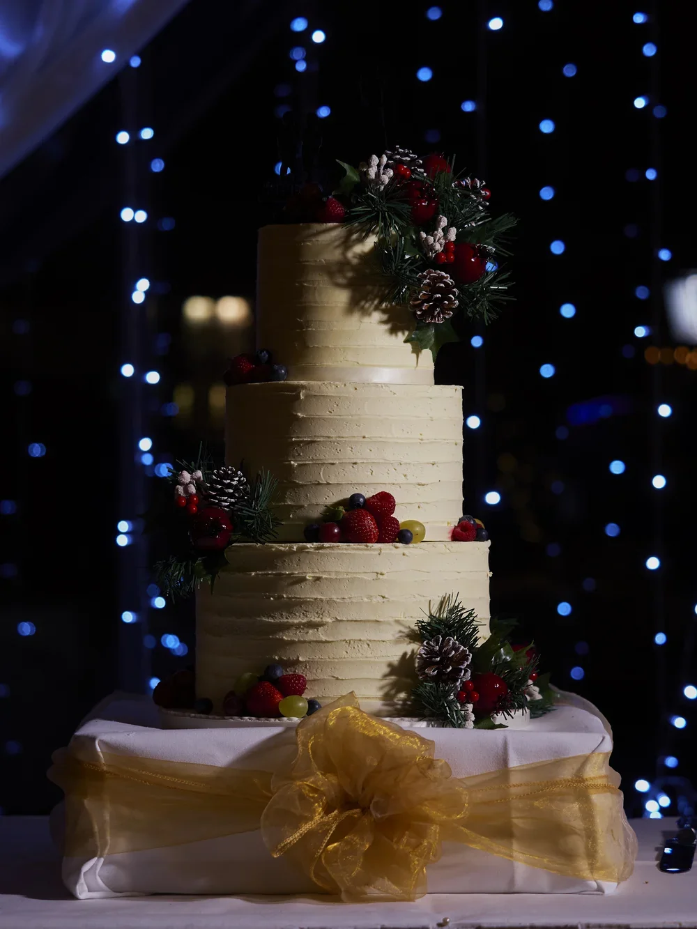 A three-tiered white frosting cake decorated with Christmas holly, berries, and pinecones, sitting on a table with a gold ribbon bow, illuminated by dark background with blue string lights.