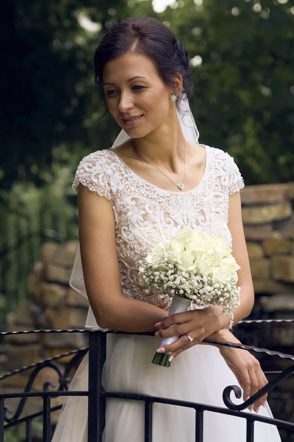 A bride in a white wedding dress with lace details on the top, holding a bouquet of white roses and baby's breath, standing outdoors near a black iron fence with greenery in the background.