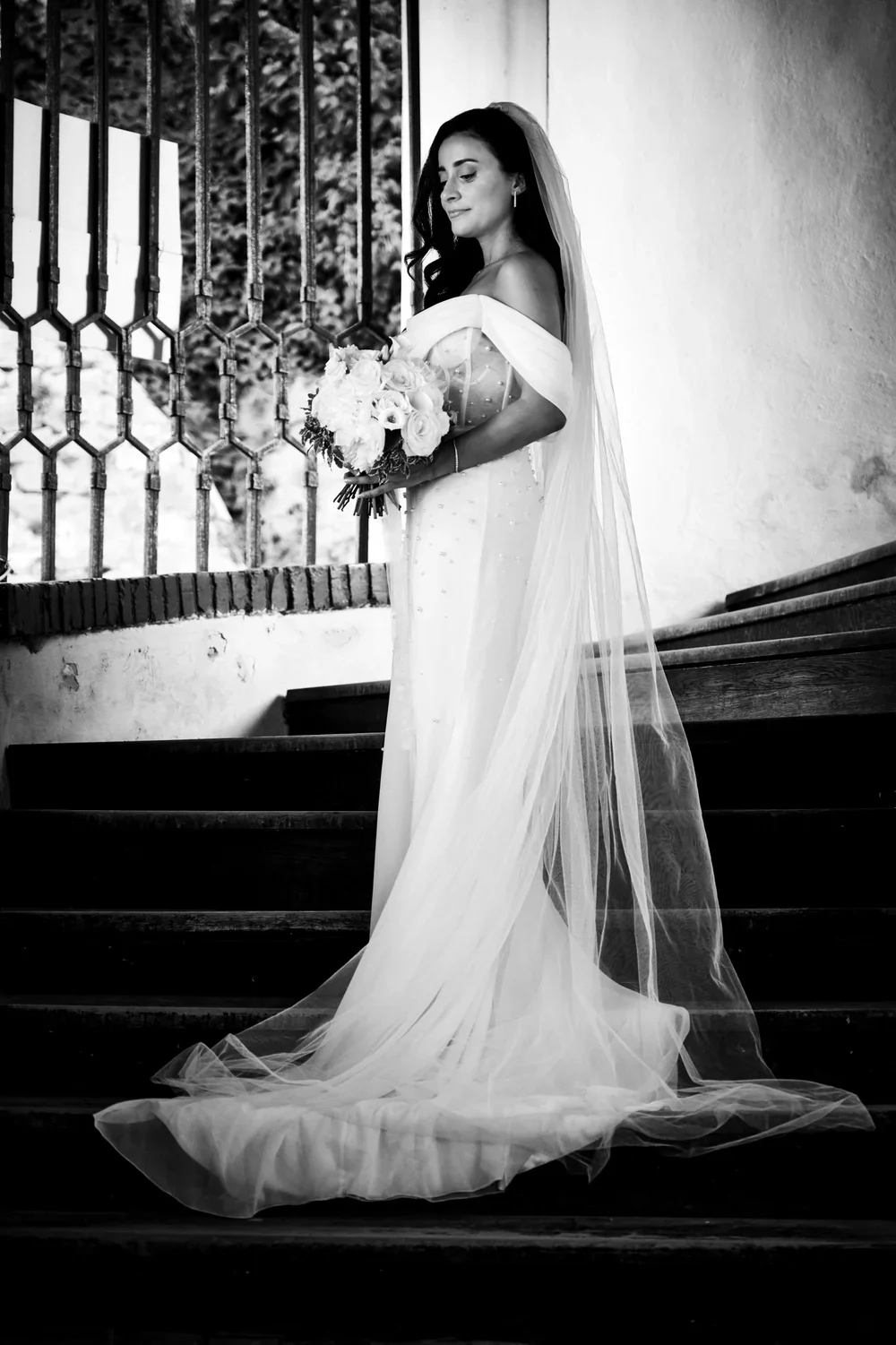 Black and white photo of a bride in a wedding dress holding a bouquet, standing on stairs near a wrought iron window.