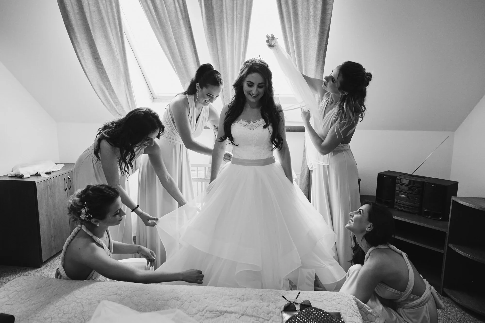 Black and white photo of a bride and her bridesmaids preparing for a wedding, with some helping her with her gown while others look on, in a room with a sloped ceiling, curtains, and a window