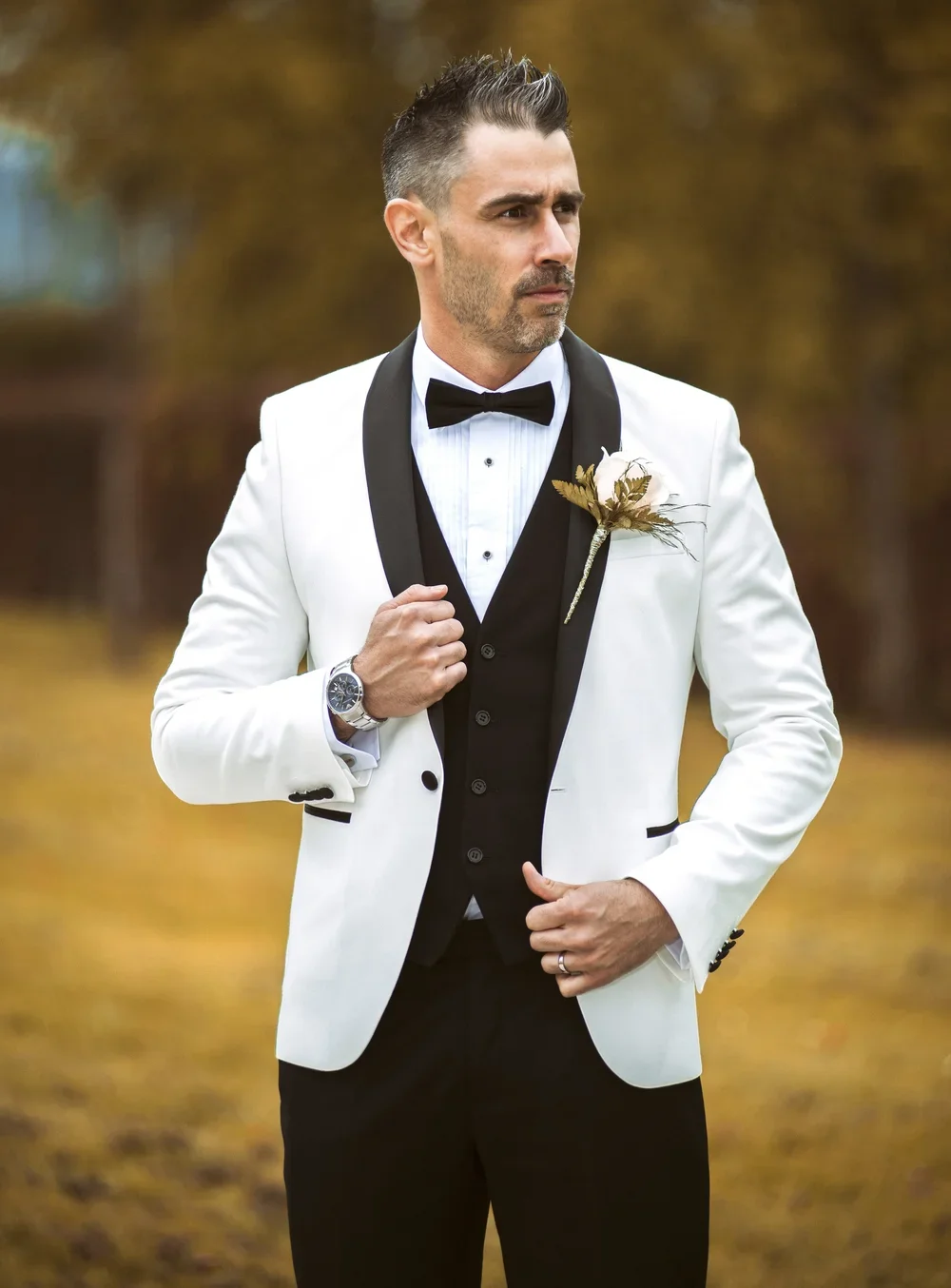 Man in a white tuxedo jacket, black vest, and bow tie, standing outdoors with fall foliage in the background, adjusting his jacket.
