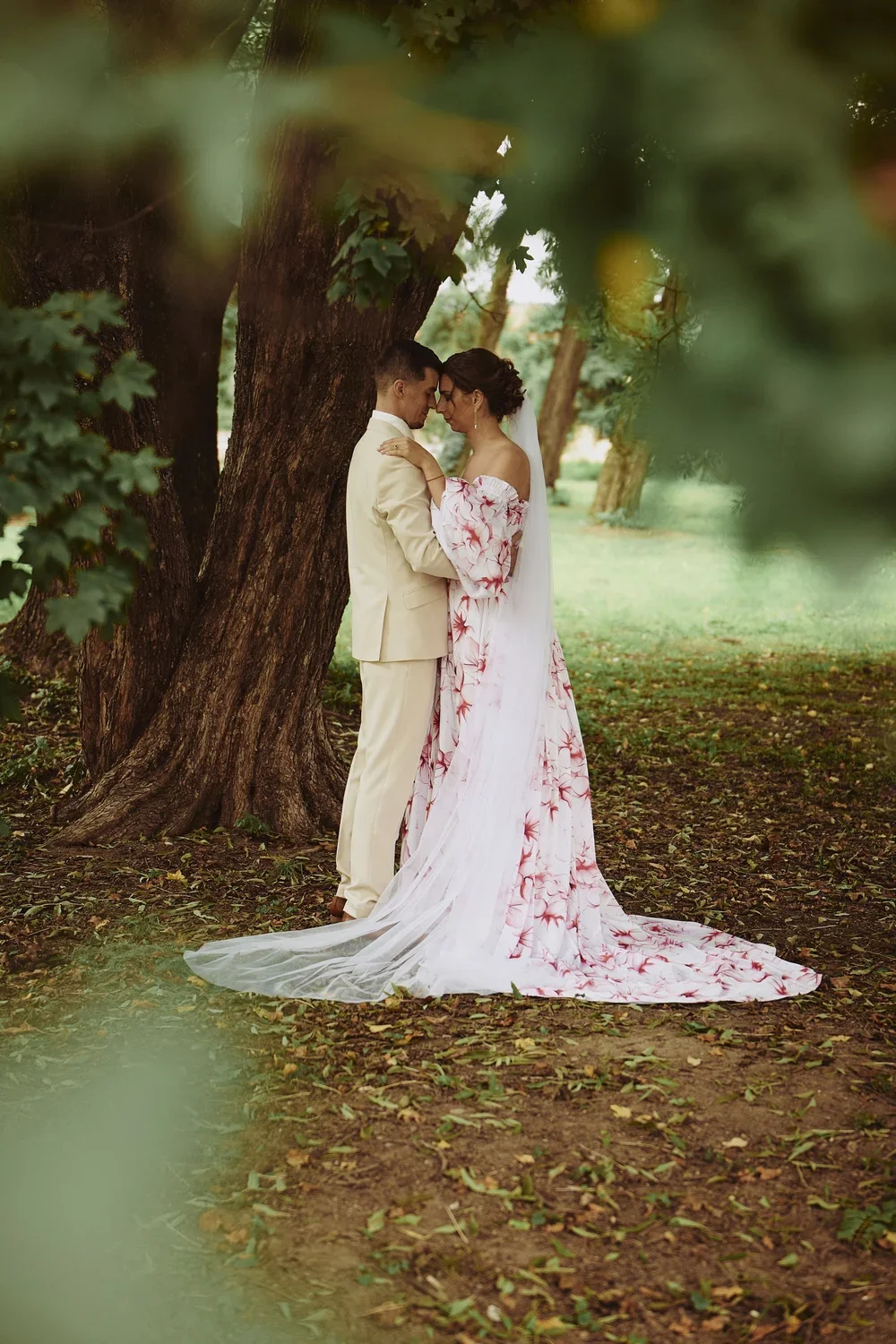 A bride and groom stand close to each other under a tree, touching foreheads in a romantic moment. The bride wears a white floral off-shoulder gown with a long veil, and the groom is dressed in a cream-colored suit. They are outdoors in a lush, green