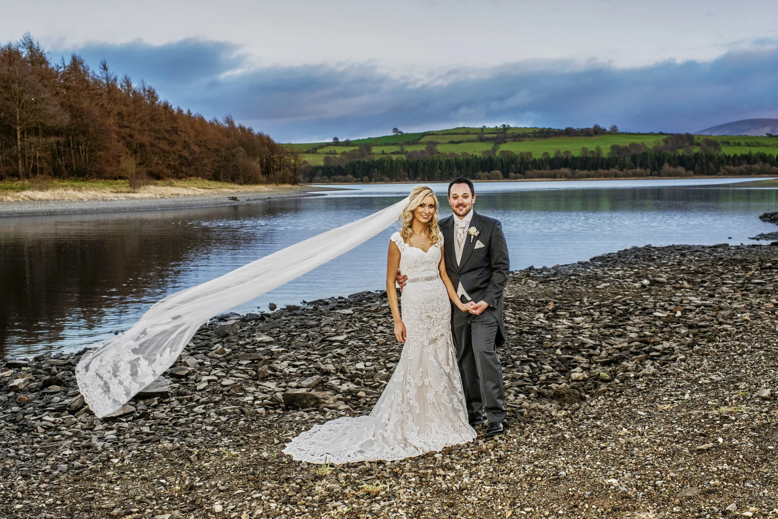 A bride and groom standing together on a rocky riverbank, with a scenic landscape of water, trees, and rolling green hills in the background, during their wedding day.