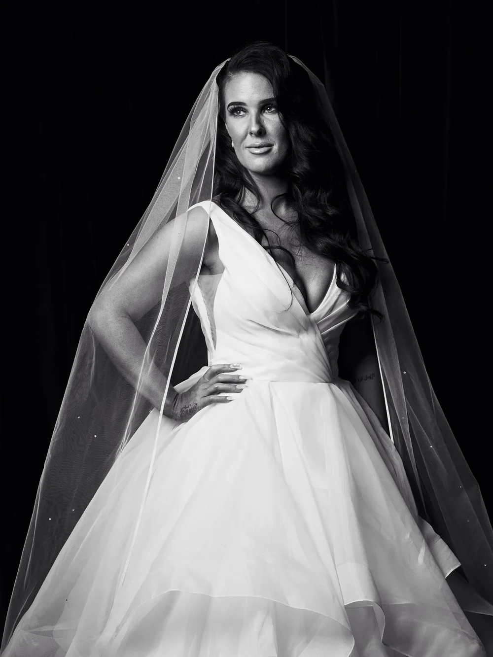 A woman with long dark hair wearing a wedding dress and veil, standing against a black background.
