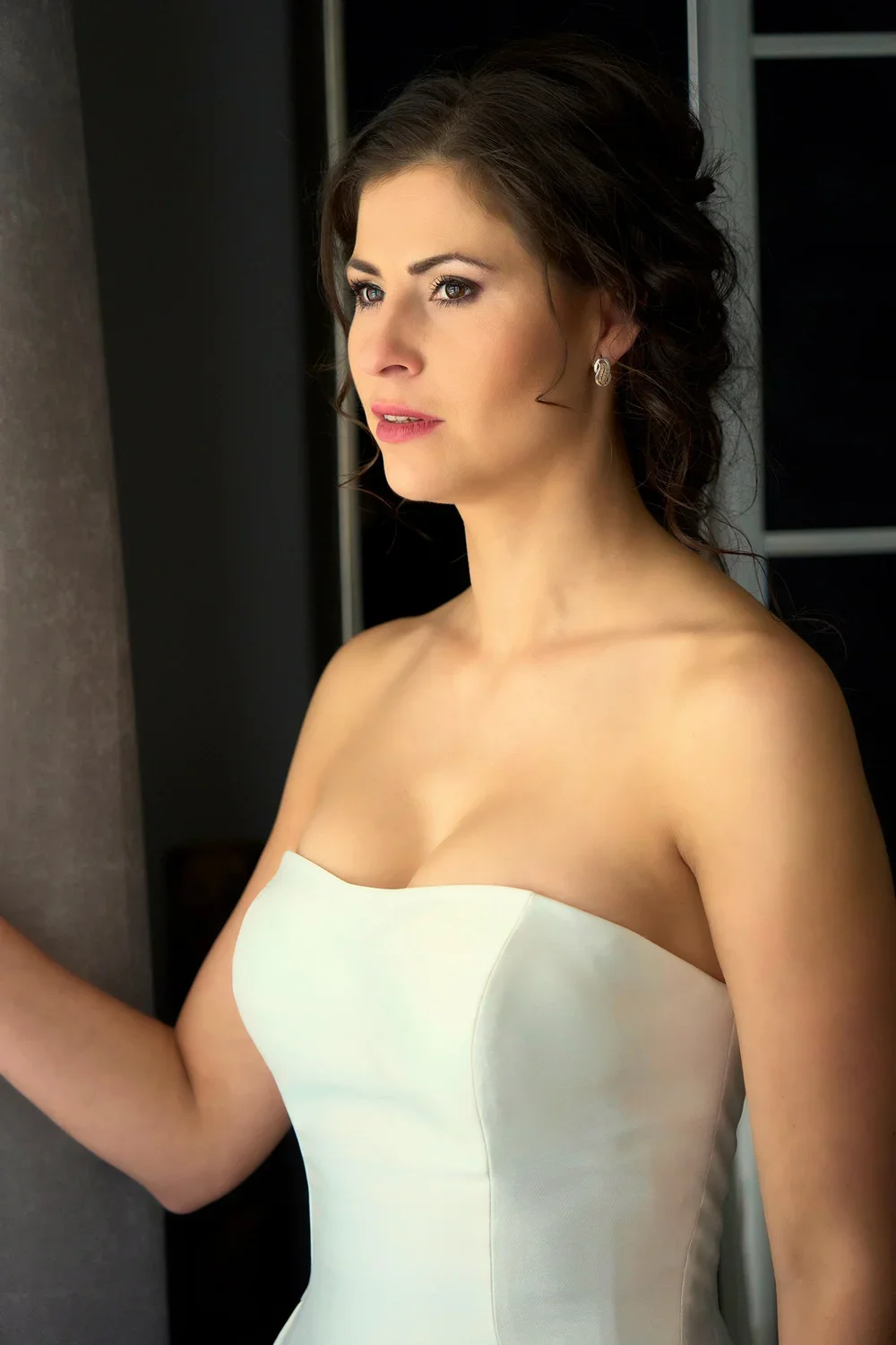 A woman in a strapless white dress with dark, wavy hair and earrings is looking to the side against a dark background.
