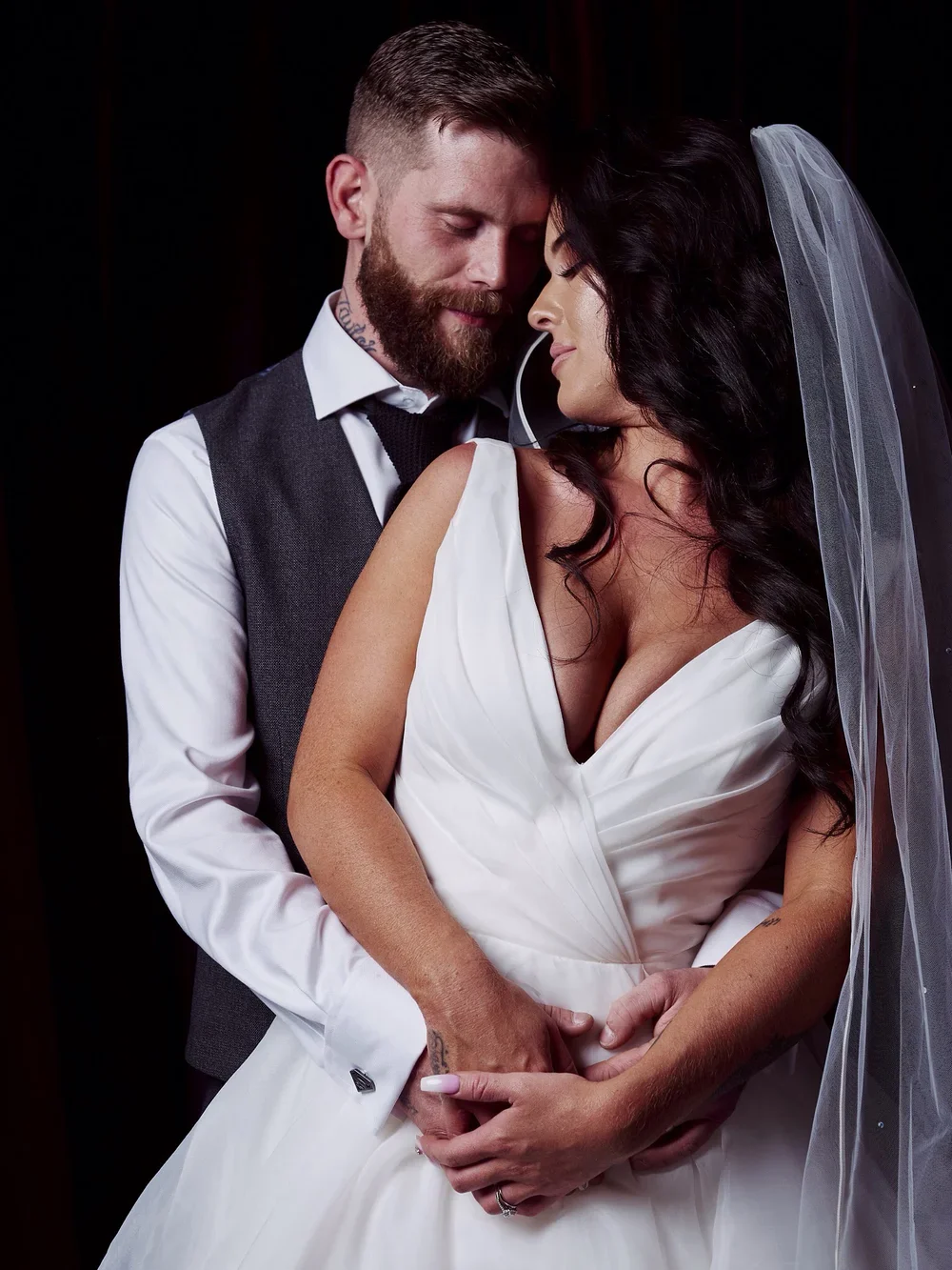 A bride and groom at their wedding, holding each other with eyes closed, dressed in wedding attire, against a dark background.