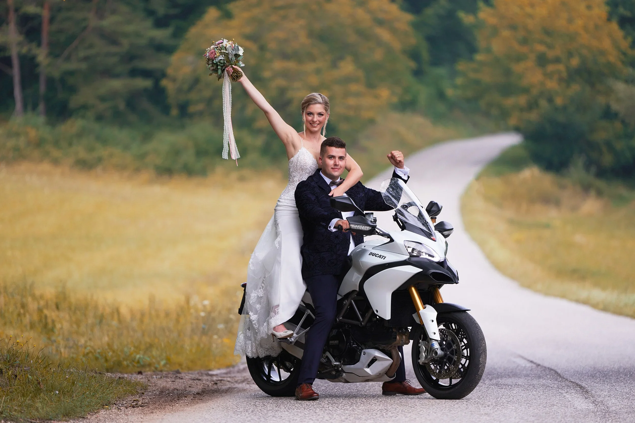 A bride and groom on a motorcycle in a rural outdoor setting with trees and a dirt road in the background. The bride holds a bouquet and raises her arm, smiling, while the groom sits on the motorcycle wearing a black suit.