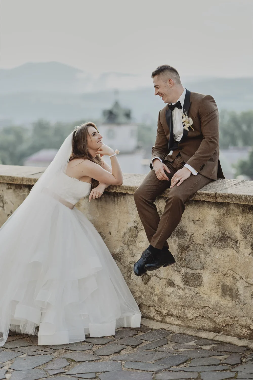 Bride and groom sharing a joyful moment on a balcony with scenic background, the bride leaning on a stone ledge, wearing a white wedding gown and veil, and the groom sitting on the ledge in a brown tuxedo with a boutonnière, smiling at each other.