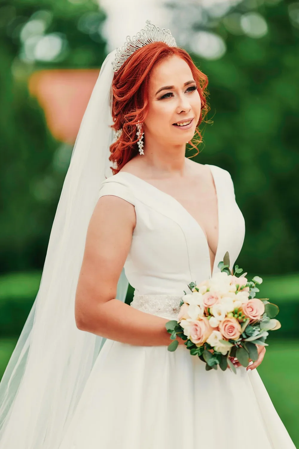A bride with red hair wearing a white wedding dress, tiara, and earrings, holding a bouquet of pink and white roses with greenery outdoors.