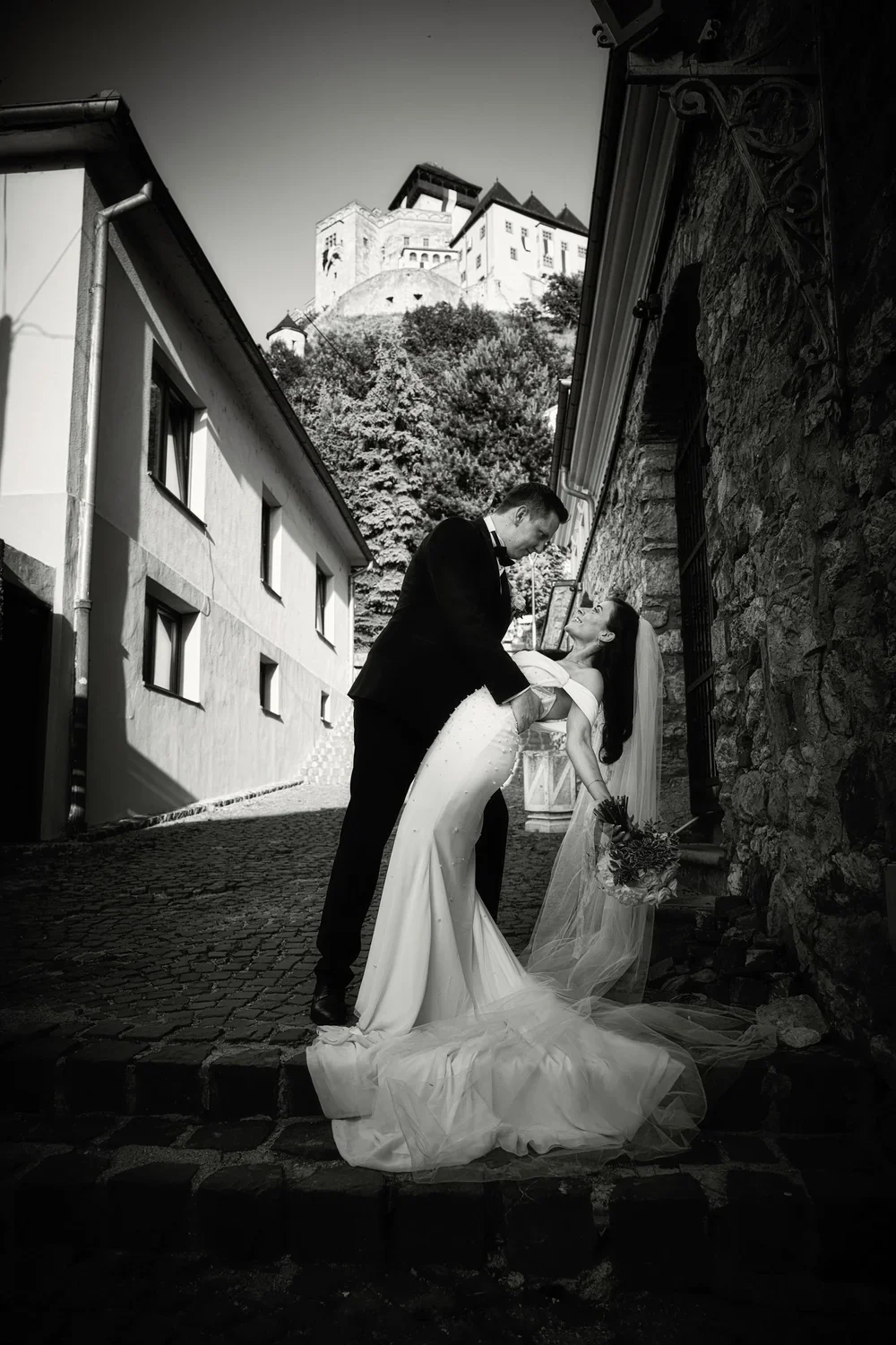 Čiernobiela svadobná fotografia: Ženích drží nevestu v romantickom záklone na kamennej dlažbe, zatiaľ čo v pozadí sa na kopci týči hrad.