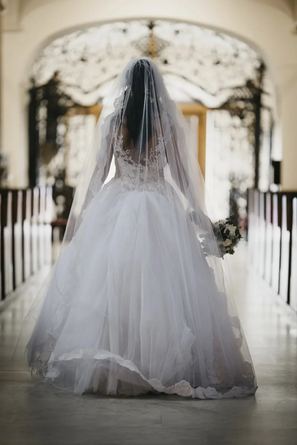 Bridal gown with lace bodice and full tulle skirt, woman standing in a church aisle wearing a veil and holding a bouquet.
