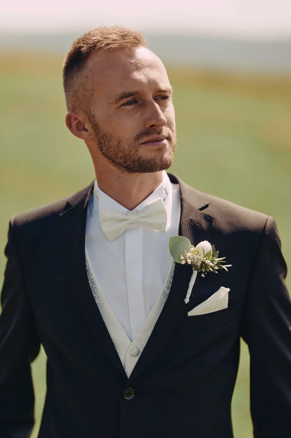 A man in a tuxedo with a bow tie and boutonniere standing outdoors on a sunny day.