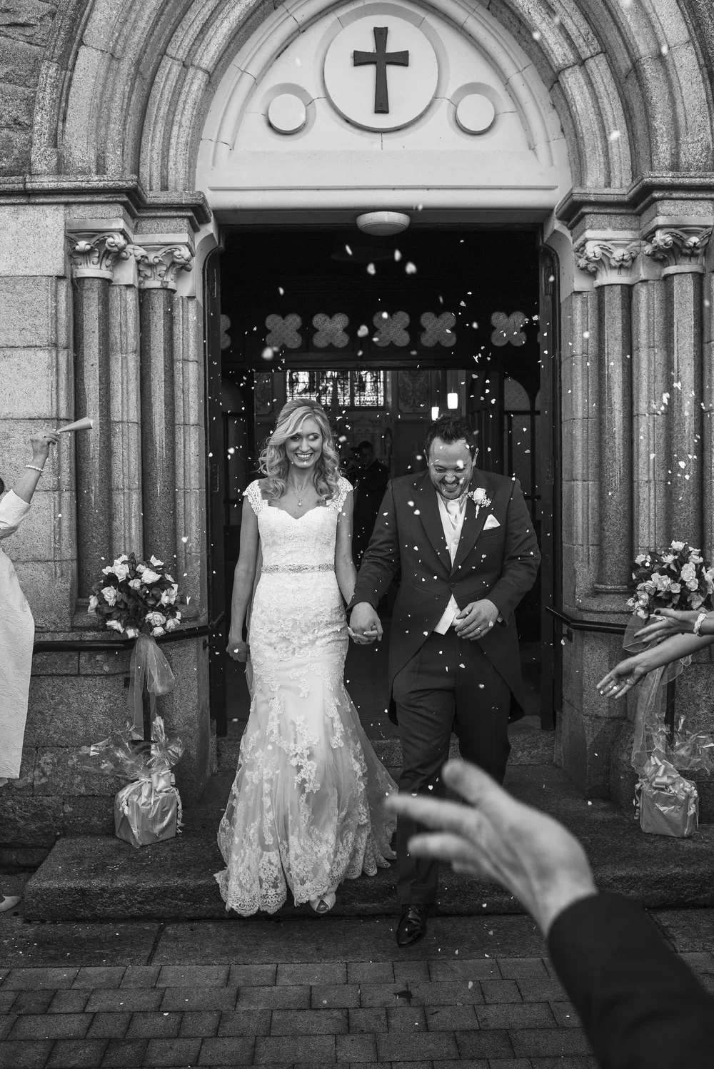 A newlywed couple holding hands and smiling as they walk out of a church, with confetti falling around them during their wedding celebration.