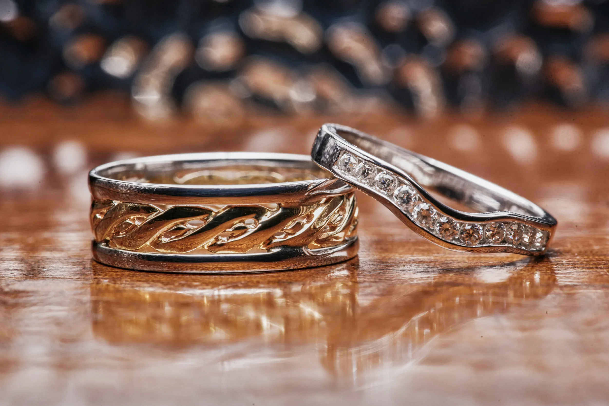 Close-up of two rings on a wooden surface, one with embedded diamonds and the other with an intricate intertwined metal design.