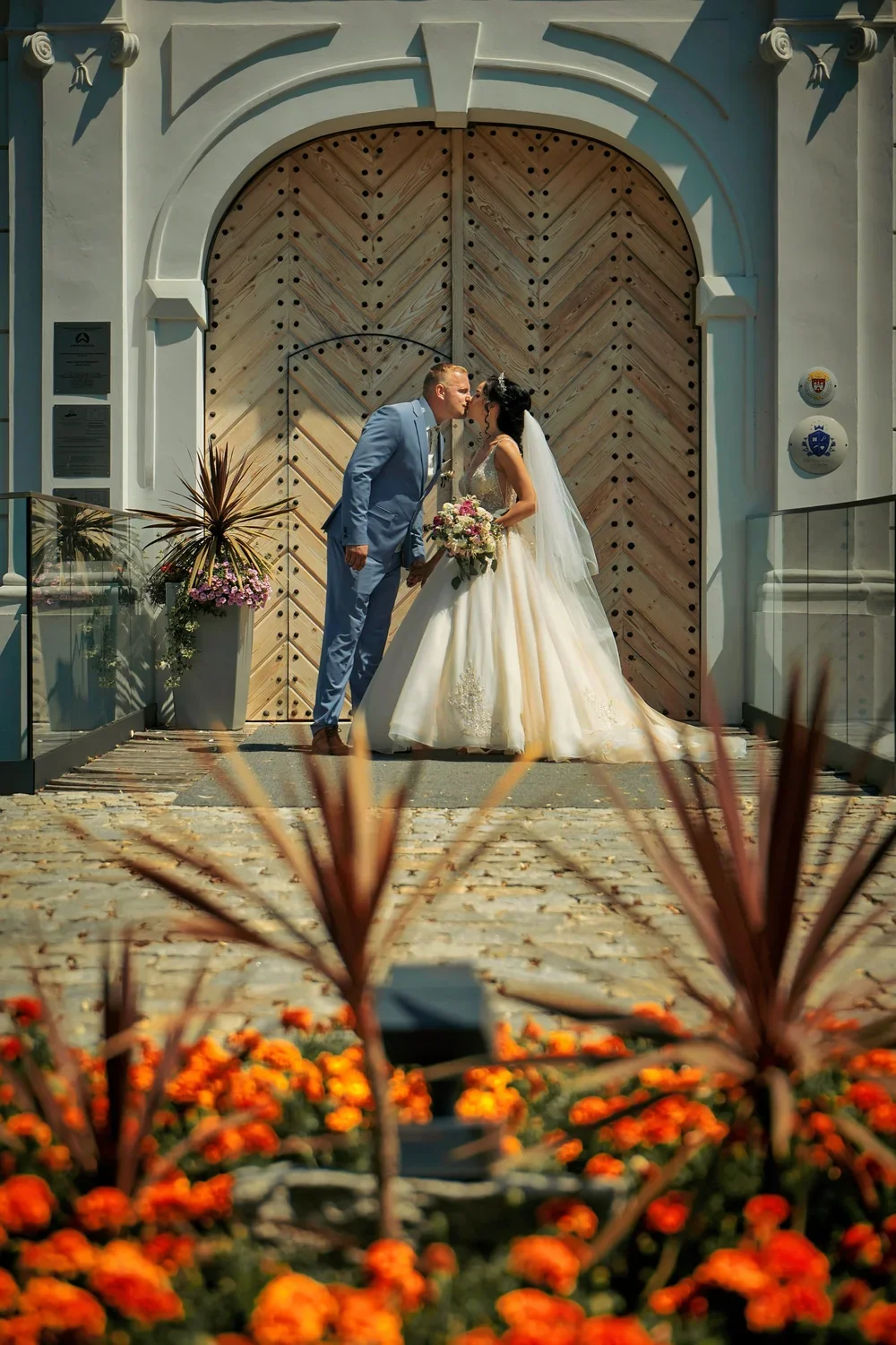 A bride and groom kiss in front of a large wooden door, surrounded by flowers and decorative plants.