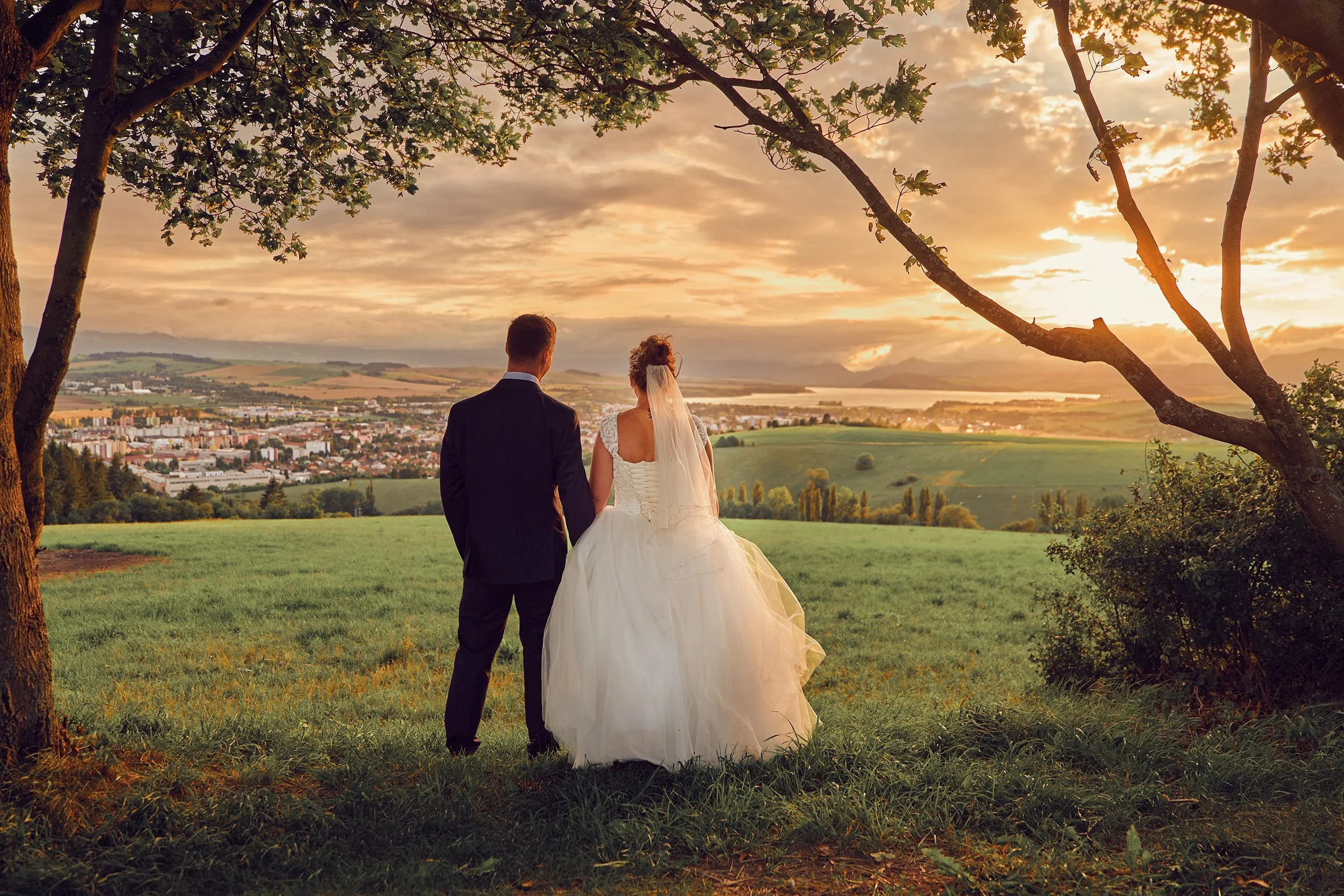 A bride and groom standing together outdoors during sunset, facing a city landscape with rolling hills, trees, and a body of water in the distance. They are framed by tree branches and leaves.