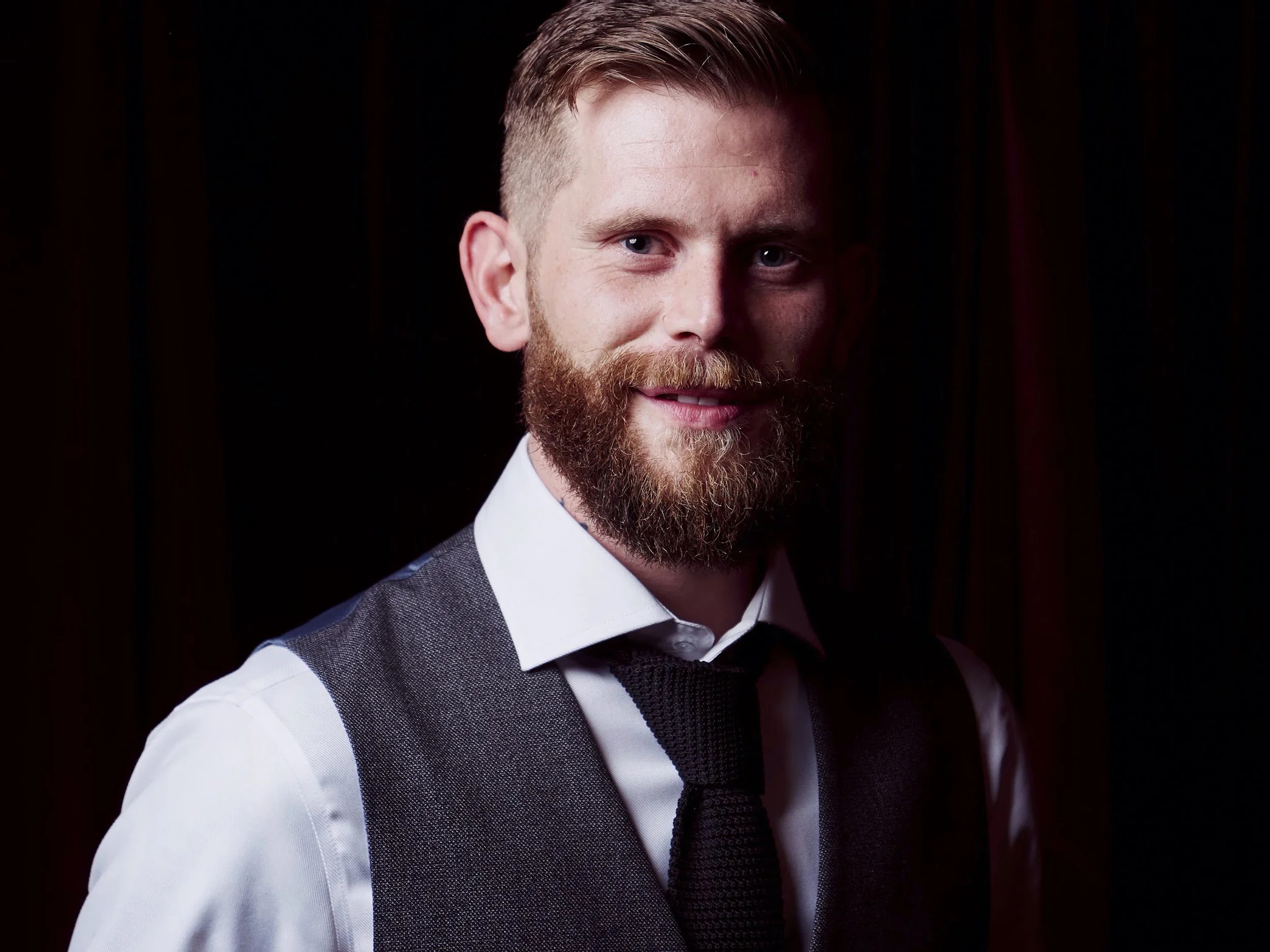 Close-up portrait of a man with red hair, a full beard, wearing a white shirt and a gray vest, against a dark background.