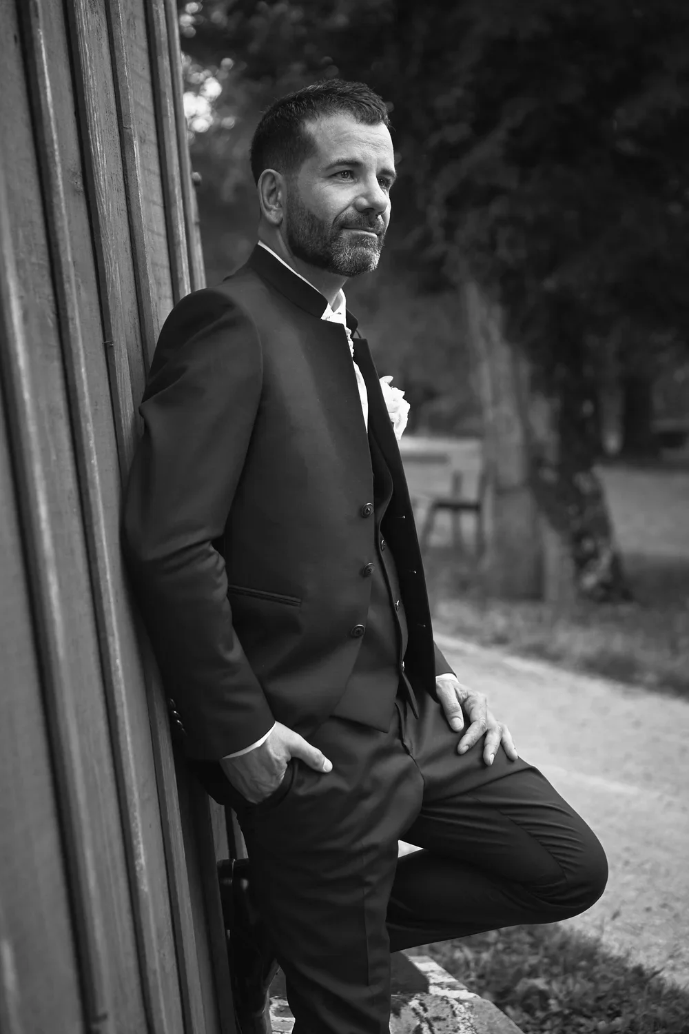 A man in a suit with a boutonniere leaning against a wooden wall outdoors, black and white photo.