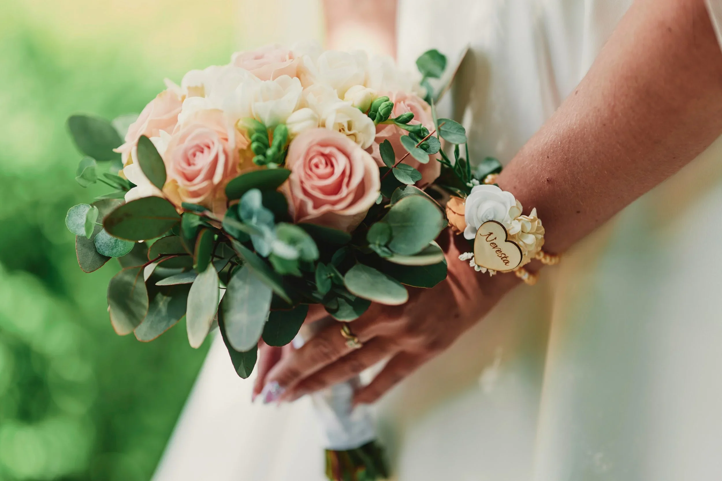 Close-up of a person holding a bouquet of pink roses, white flowers, and green foliage, with a small handmade heart charm attached to the bouquet.