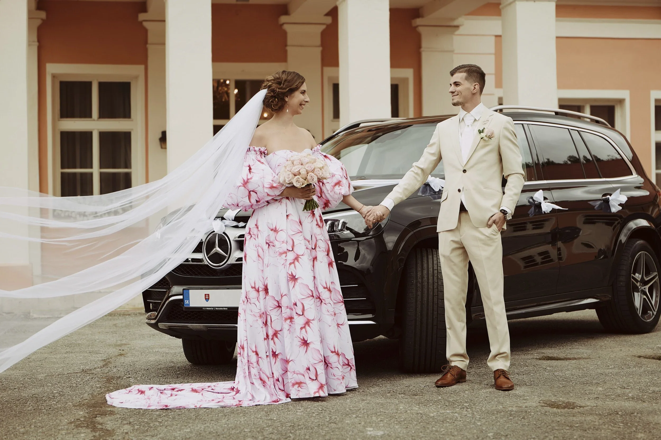 A bride and groom holding hands in front of a black Mercedes-Benz decorated with white ribbons, standing outside a peach-colored building with columns.