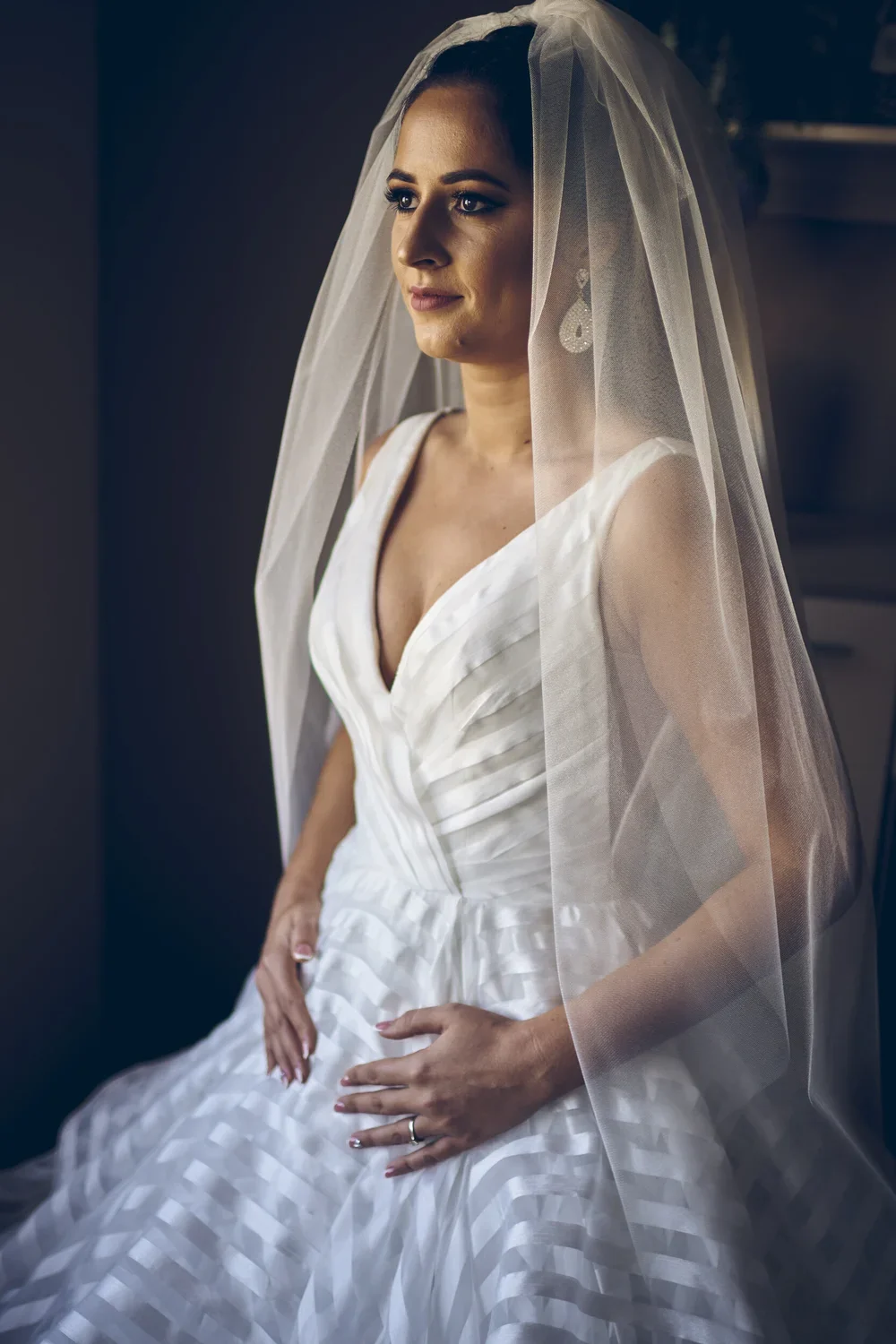 A bride in a satin wedding dress with a deep V neckline, wearing a veil and earrings, sitting indoors with a calm expression.