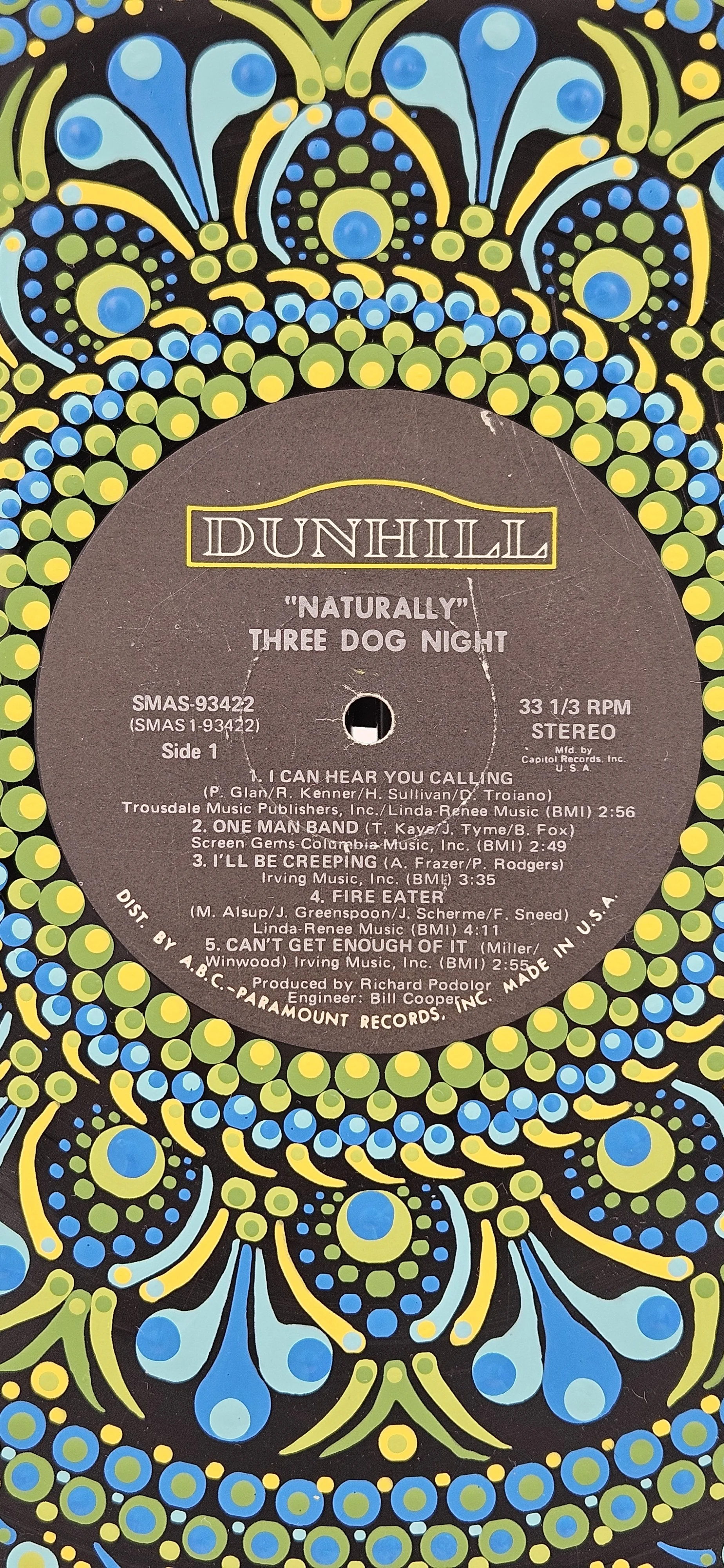 Three Dog Night Naturally hand-painted vinyl record art displayed on a tabletop stand in a room with blue and green accent décor