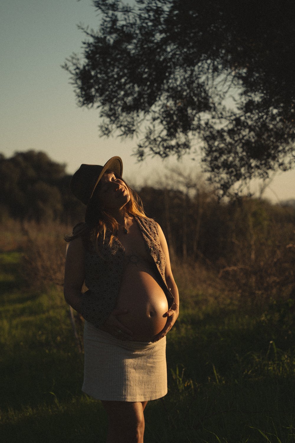 Photo of a pregnant women in the soft evening sunlight. She holds her hands around her belly. The picture is soft and serene. The photograph is taken in nature.