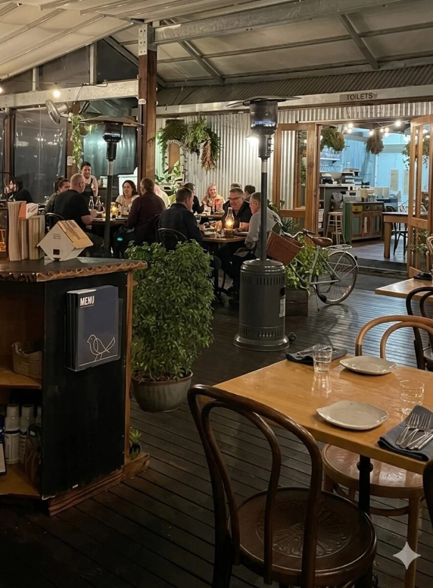 A cozy indoor restaurant or cafe with wooden furniture, potted plants, and a group of people dining at a long table. There are heaters and decorative hanging plants, and a bicycle outdoors visible through a door.
