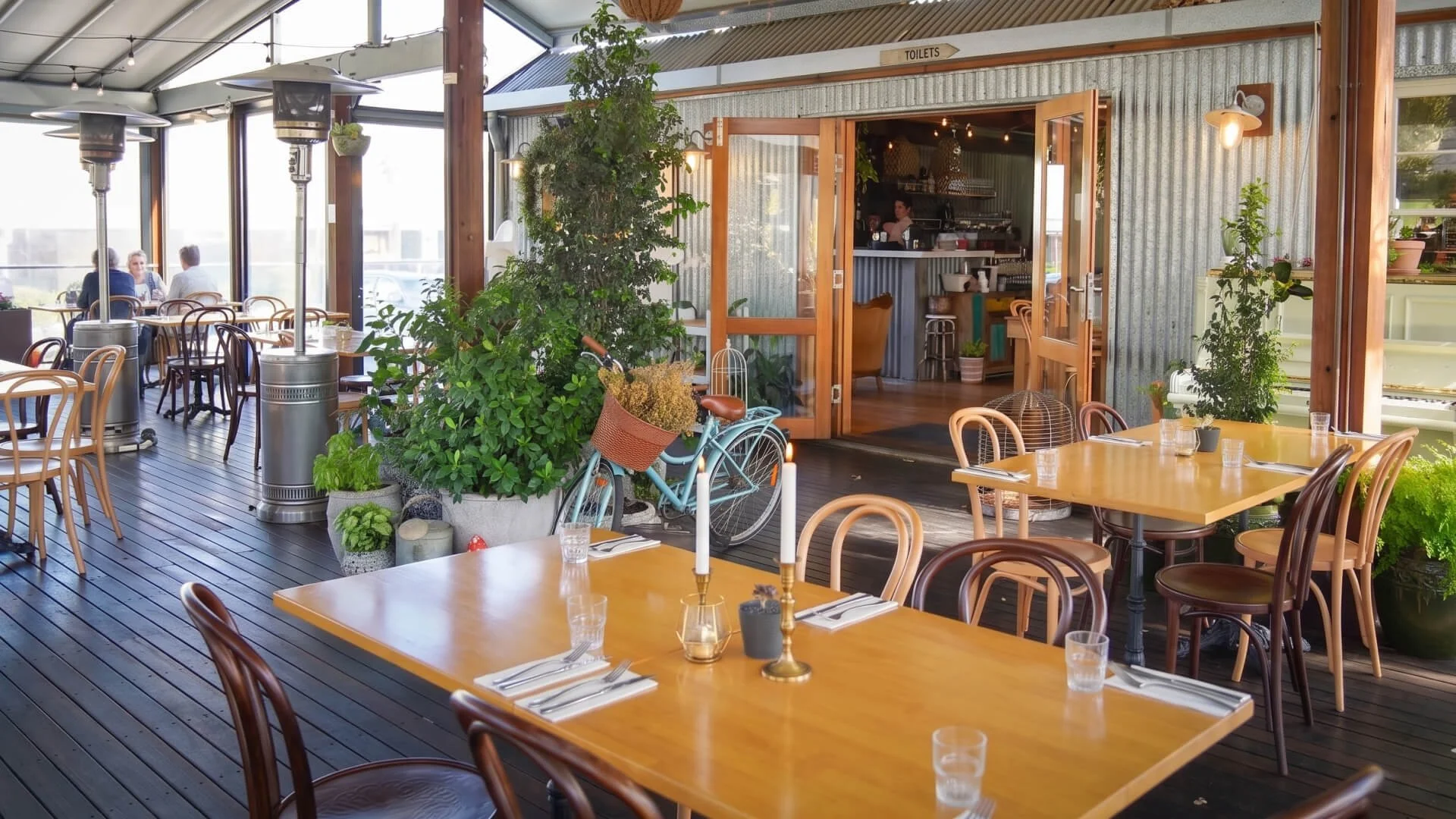 Indoor restaurant with wooden tables and chairs, potted plants, a bicycle with a basket, and large windows letting in natural light.