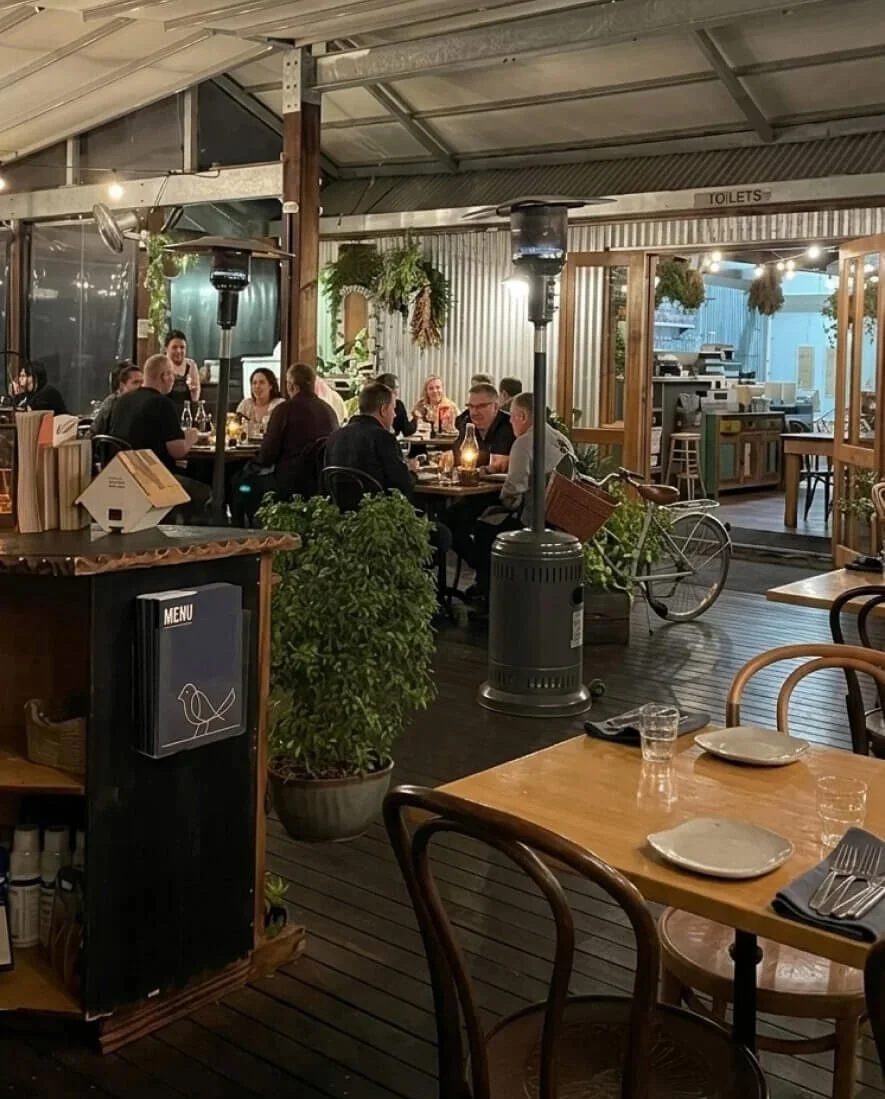 Indoor restaurant with patrons seated at tables, some with candles, potted plants, a bicycle, and a heater. The atmosphere is cozy and casual.