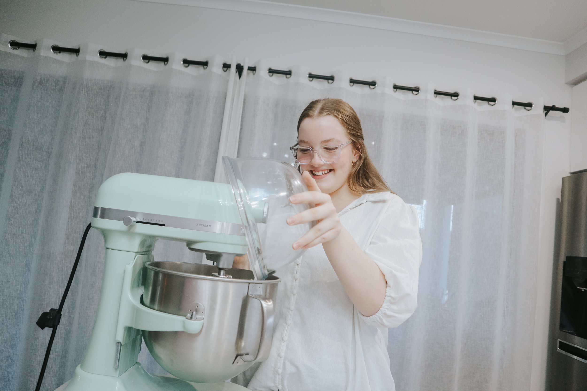 A woman smiling and looking down at a stand mixer in a bright room with light-colored curtains.