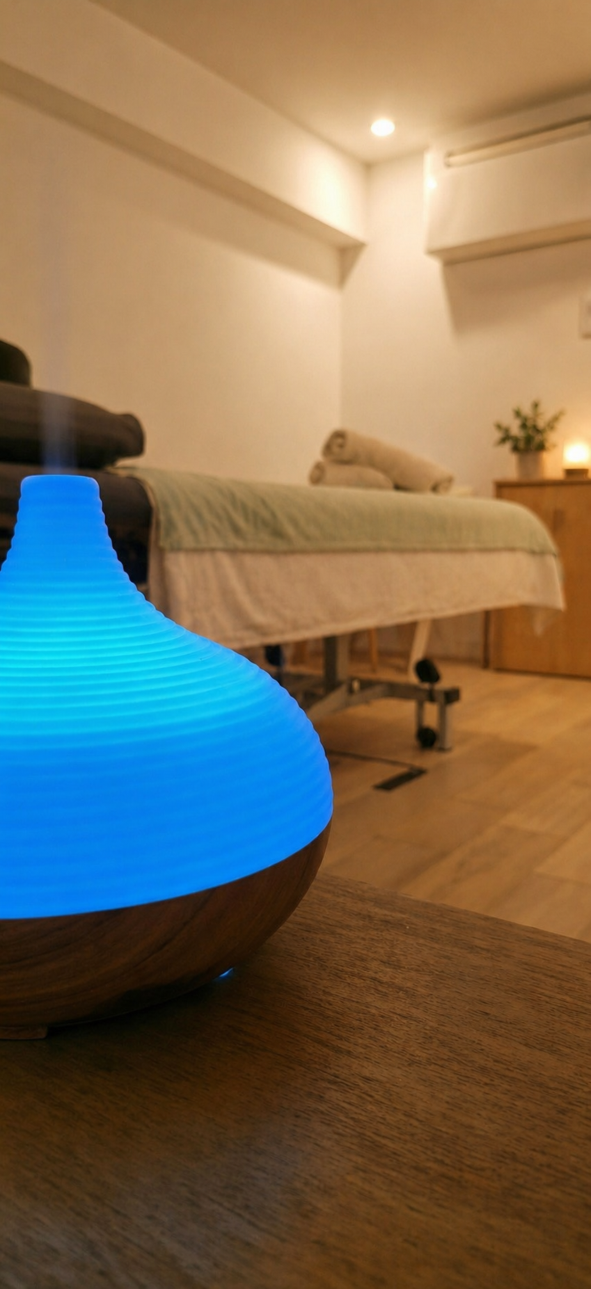 A blue LED aromatherapy diffuser on a wooden table in a room with a massage bed, towels, a potted plant, and soft lighting.