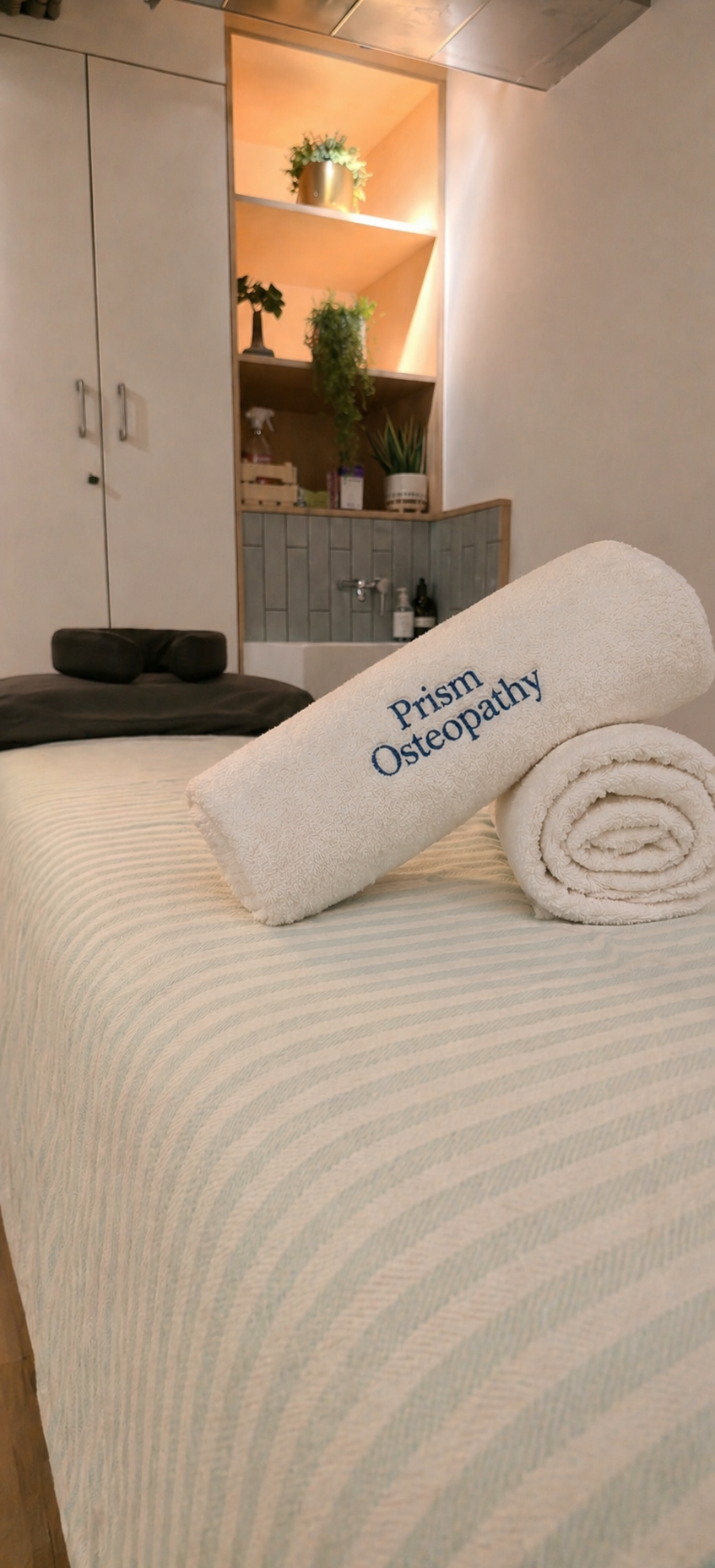 A massage table with a rolled towel embroidered with 'Prism Osteopathy' in a cozy room featuring a shelf with plants and a small sink.