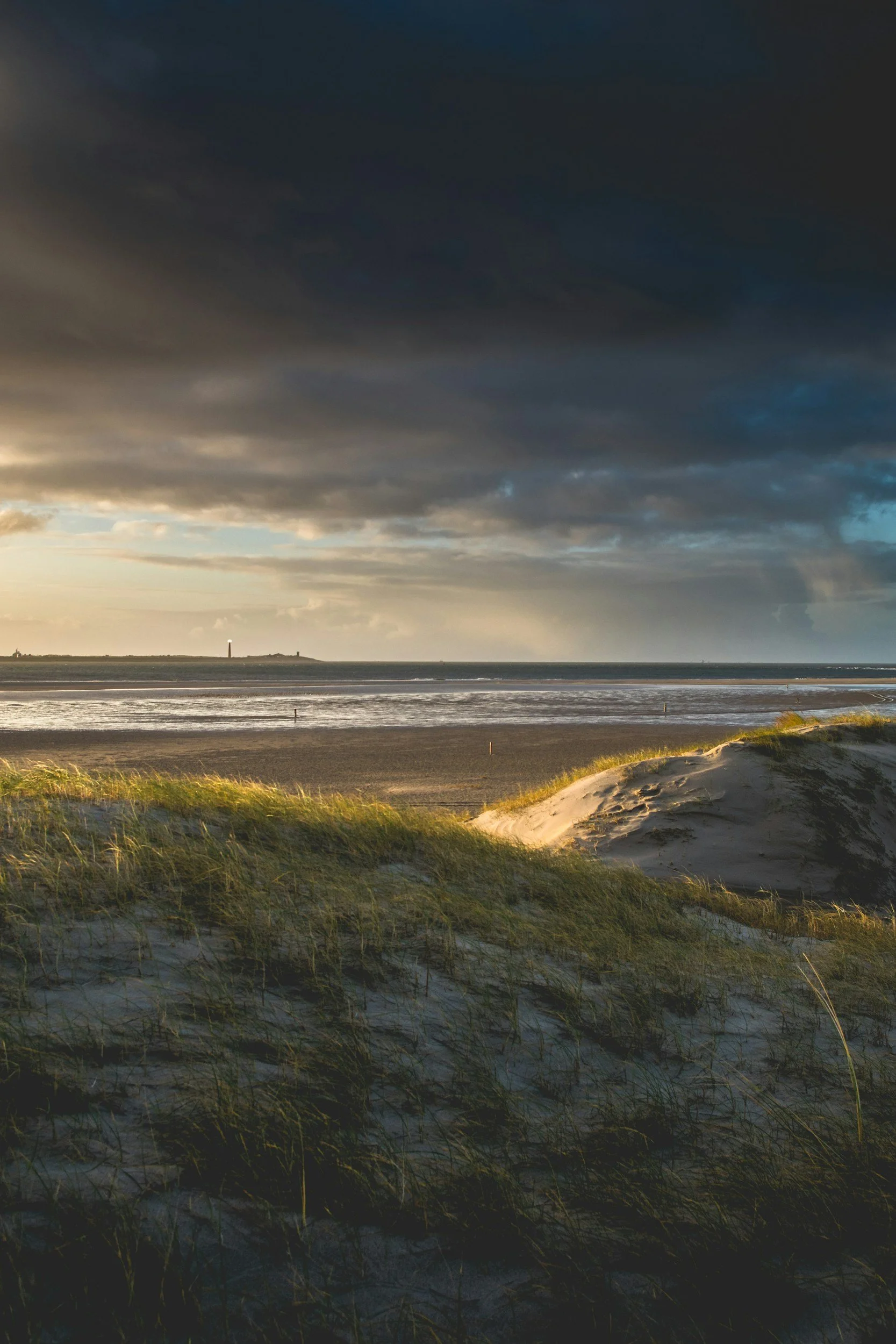 Strand bei Sonnenuntergang mit dunklen Wolken am Himmel und Dünen im Vordergrund.