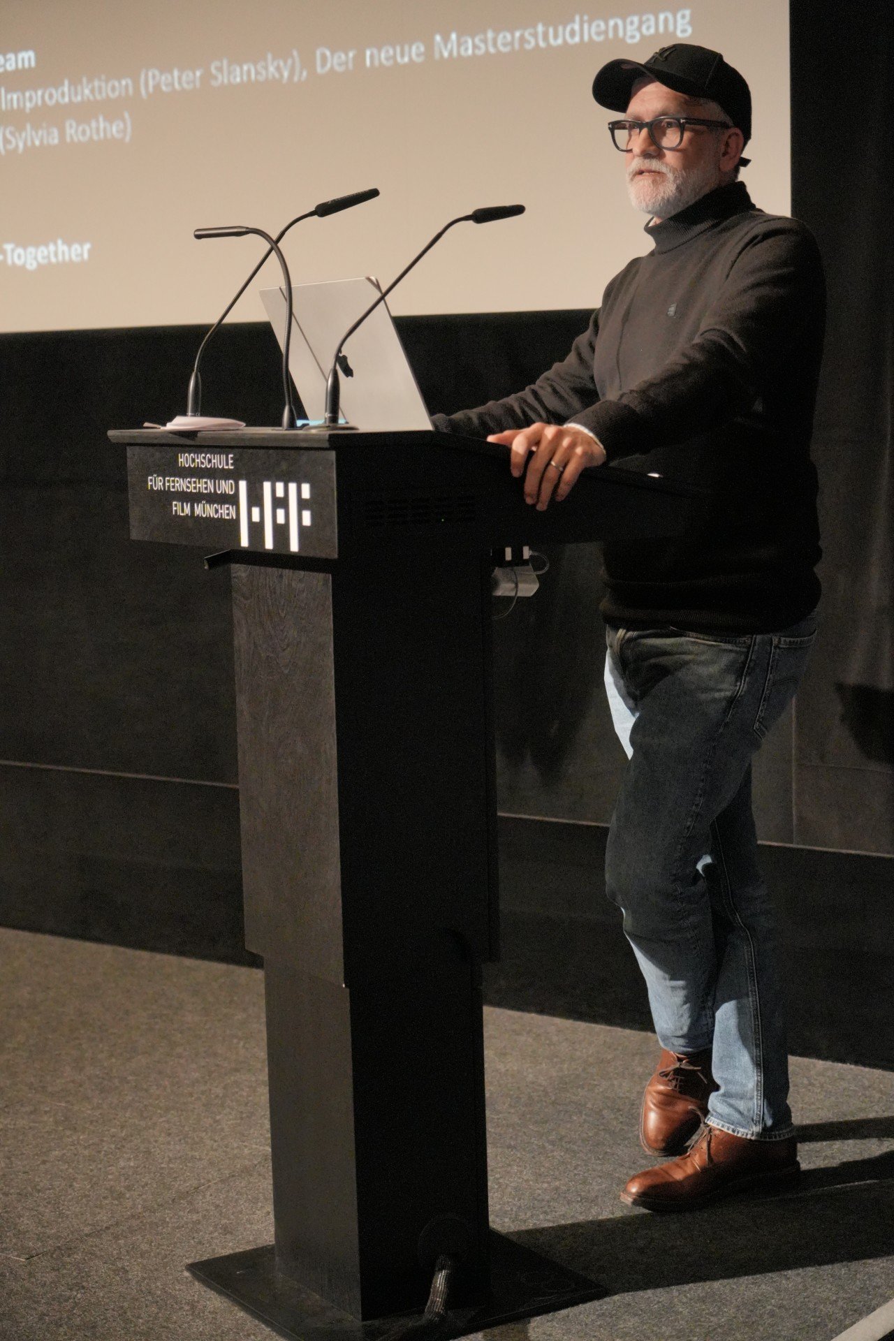 Taç Romey spricht an einem Rednerpult der Hochschule für Fernsehen und Film München (HFF) vor einer Leinwand in einem abgedunkelten Hörsaal.