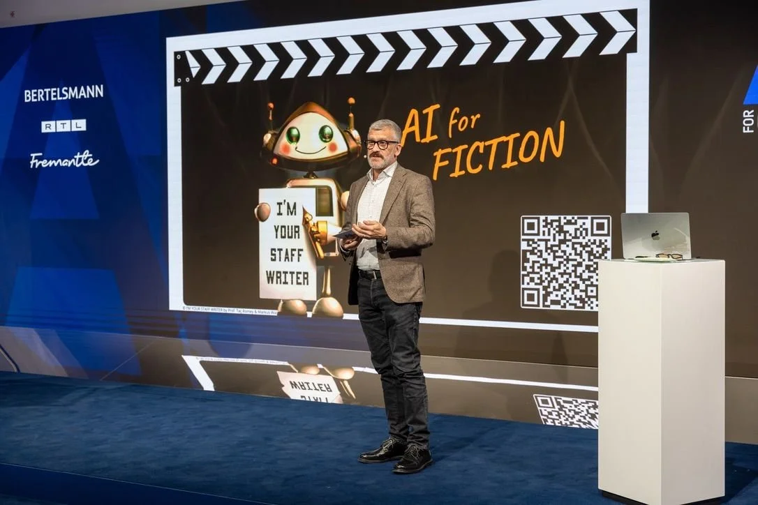 Taç Romey präsentiert auf der Bühne eines Bertelsmann/RTL/Fremantle-Events vor einer großen Leinwand mit der Aufschrift „AI for Fiction" und einer Roboterfigur mit dem Schild „I'm Your Staff Writer".